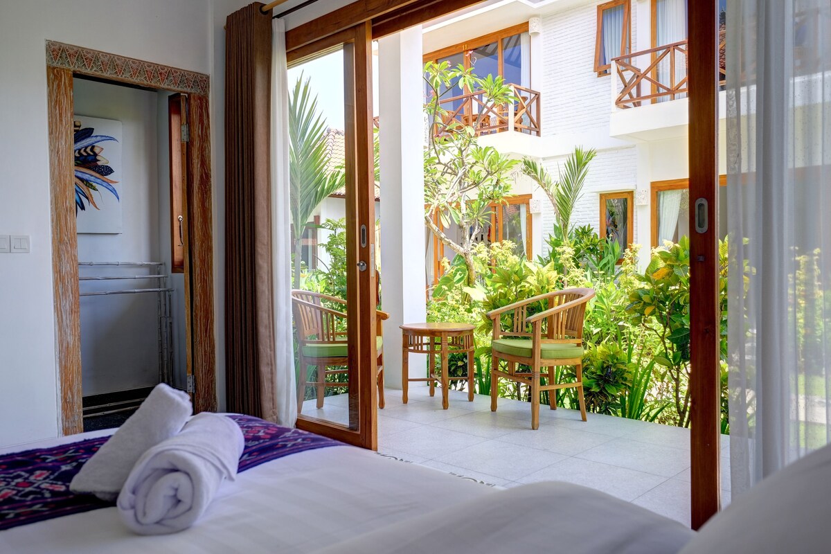 A hotel room with a bed, a wooden chair, a table, and a view of a courtyard with plants and a building.