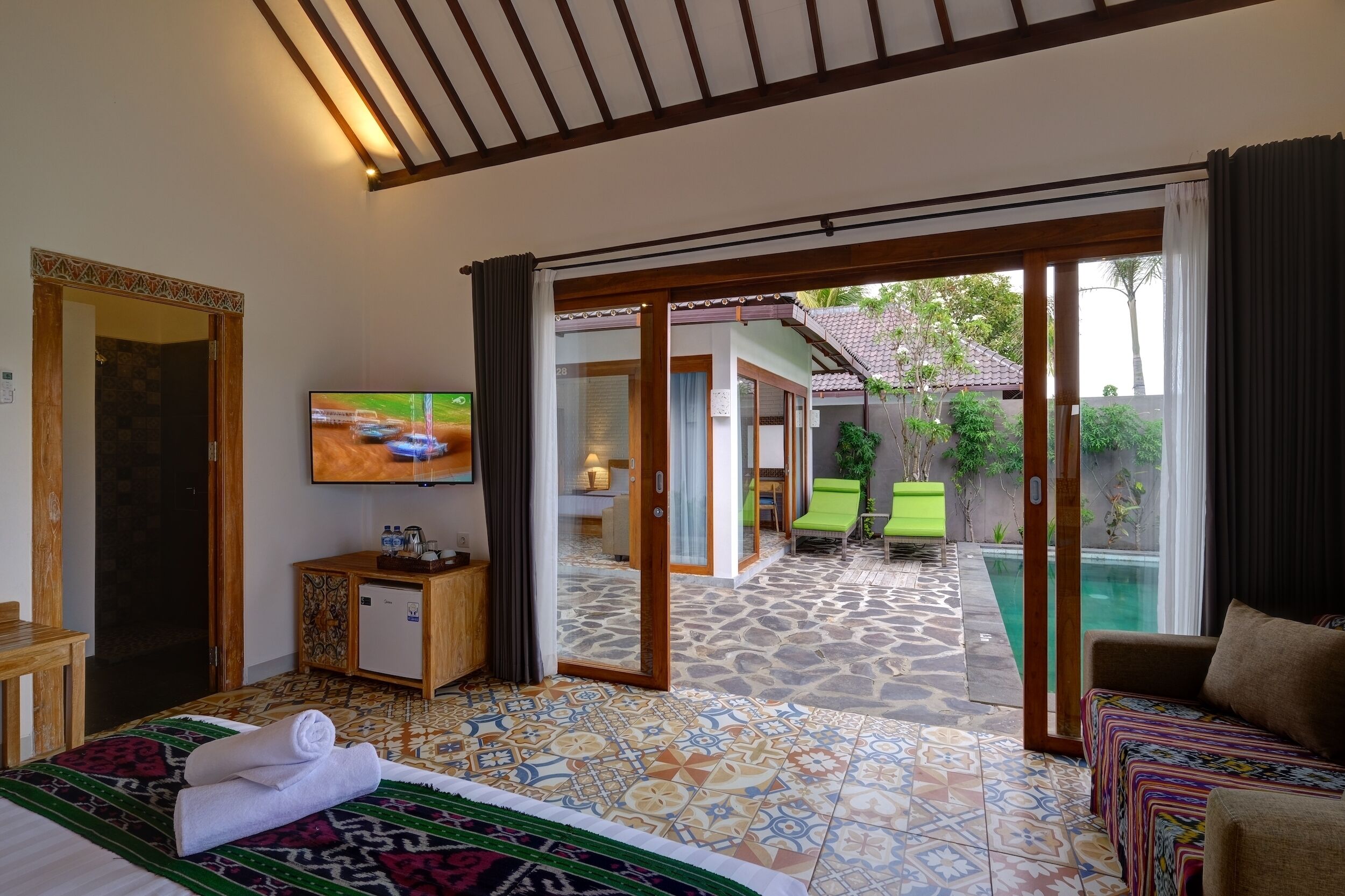 Habitación de hotel con cama, televisión, un pequeño armario y vistas a una piscina y jardín a través de una puerta corrediza de cristal.