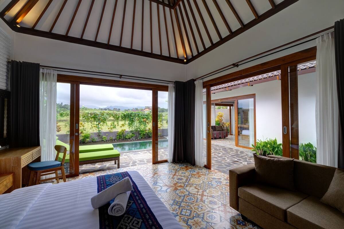 A hotel room with a bed, a sofa, a small table, and a chair. There is a large window with a view of greenery and a pool.