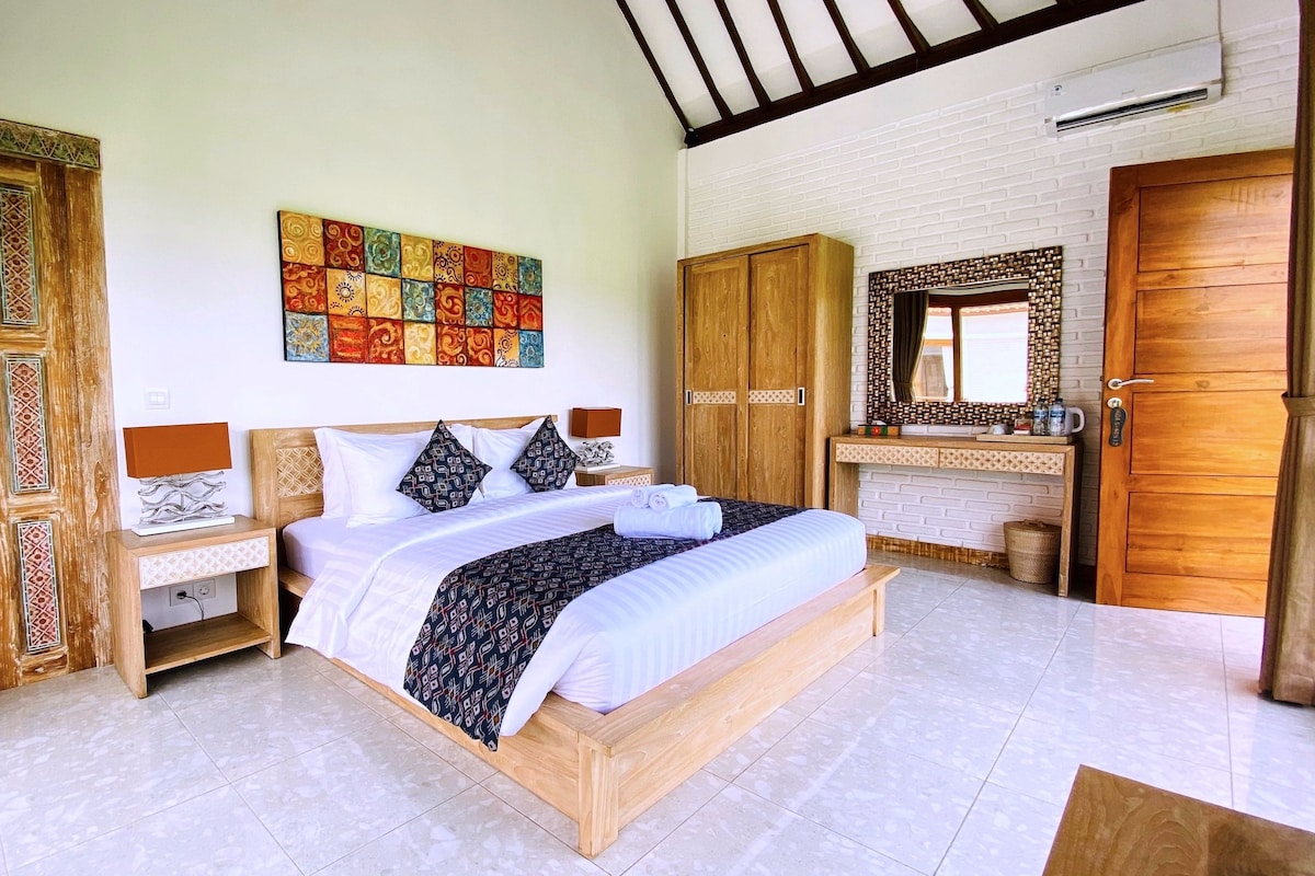 A hotel room with a large bed, wooden wardrobe, a bathroom with a mirror, and a colorful artwork on the wall.