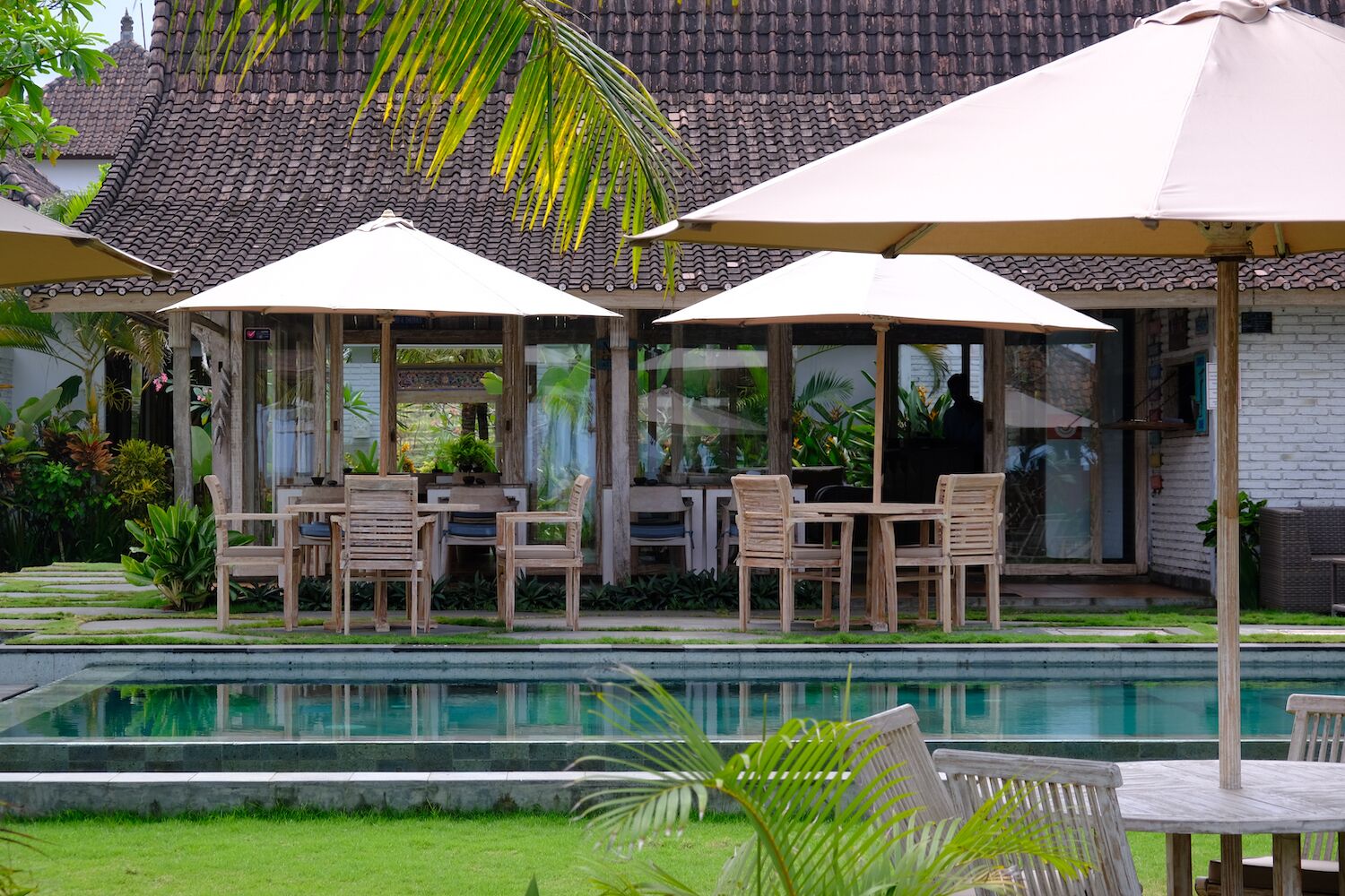 A poolside area with wooden tables and chairs, umbrellas, and a view of a traditional tiled roof building.