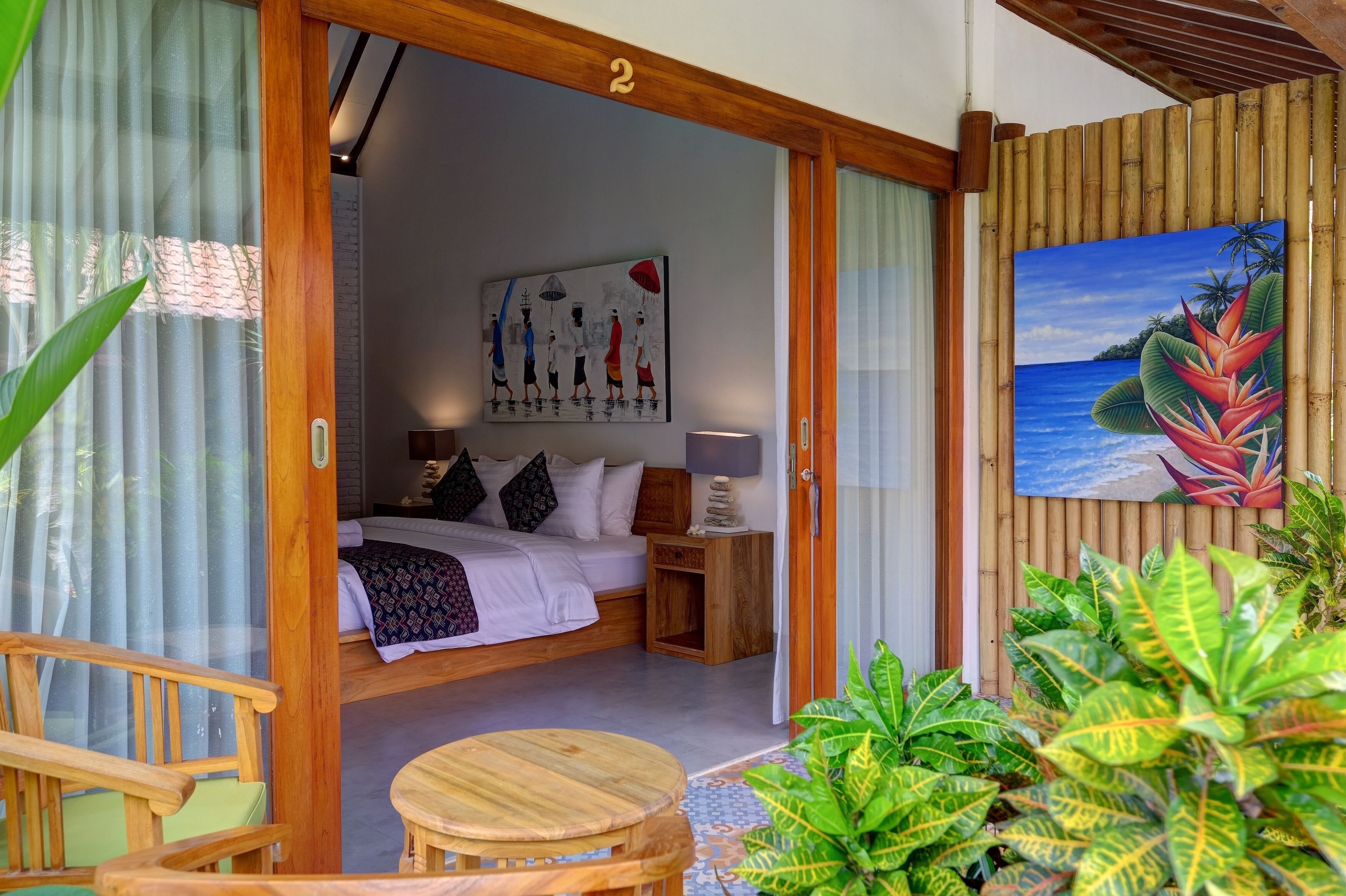 A bedroom with a bed, pillows, a nightstand, and a painting. There is a wooden door leading to the room, and a bamboo wall.