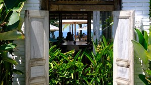 A traditional wooden gate with intricate carvings, leading to a covered outdoor seating area with tables and chairs, surrounded by lush tropical plants. - Wide Sands Beach Retreat (Mendoyo)