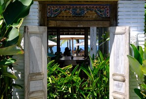 Una puerta de madera tradicional con intrincados grabados, que da paso a una zona de estar al aire libre techada con mesas y sillas, rodeada de exuberante vegetación tropical.