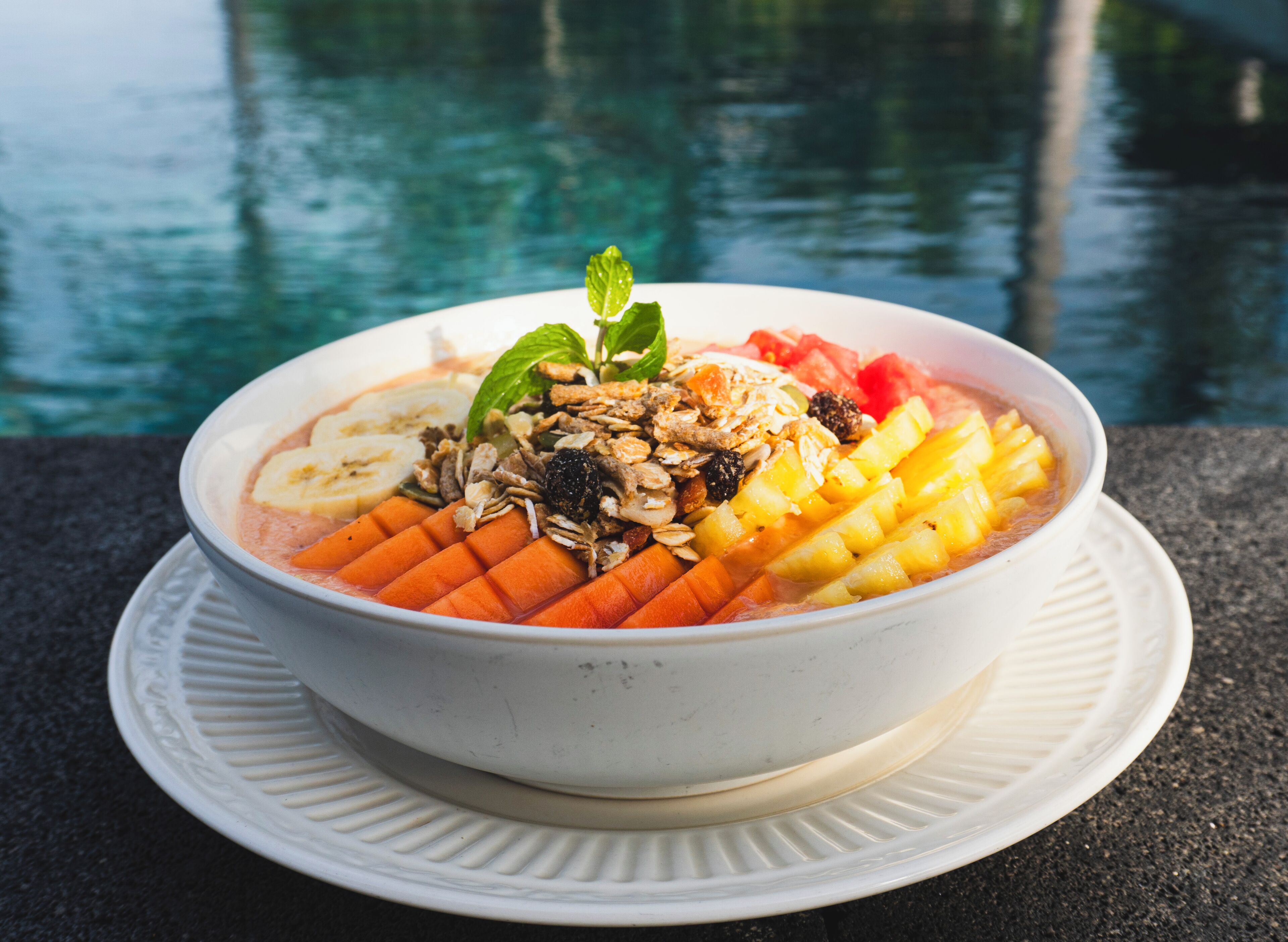 Un tazón de fruta y granola sobre un plato blanco, con una piscina al fondo.