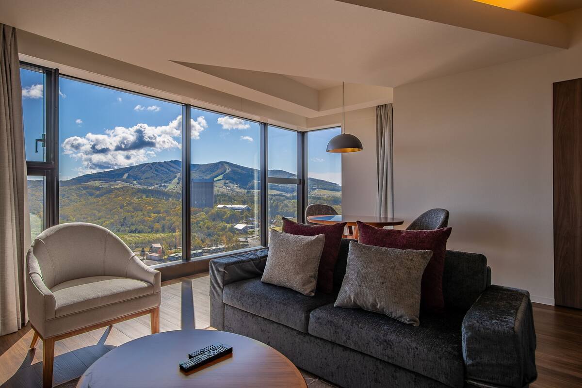 A modern living room with a sofa, armchair, and coffee table, large windows with a mountain view, and a pendant light.