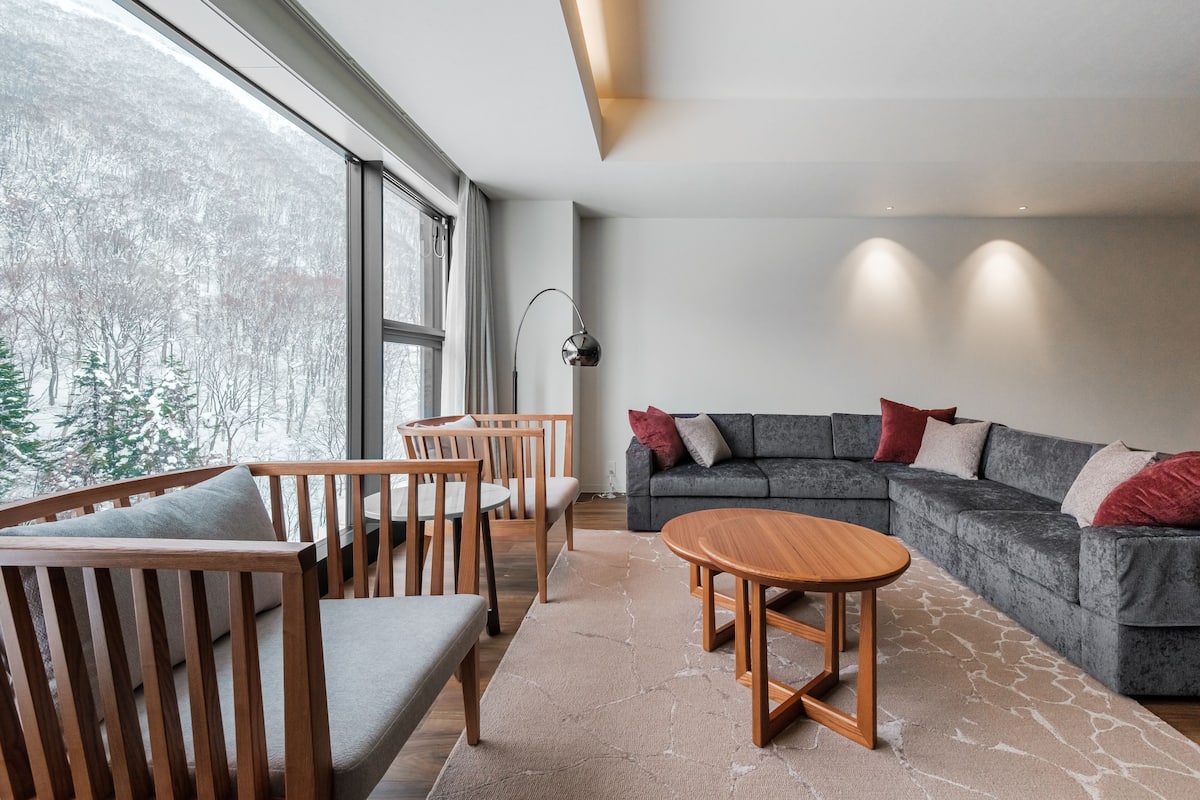 A modern living room with a grey sectional sofa, wooden chairs, a round wooden coffee table, and a large window offering a view of a snowy forest.