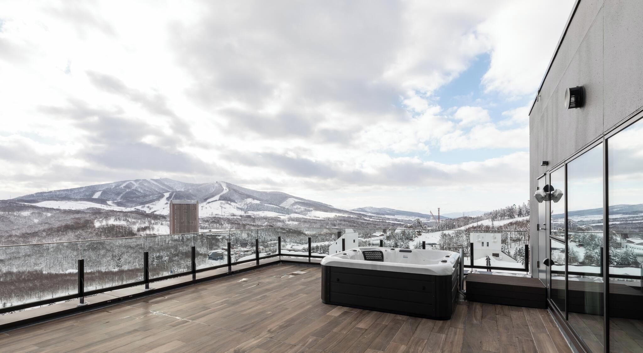 A high-rise building with a wooden deck, a black outdoor seating area, and a view of snow-covered mountains.