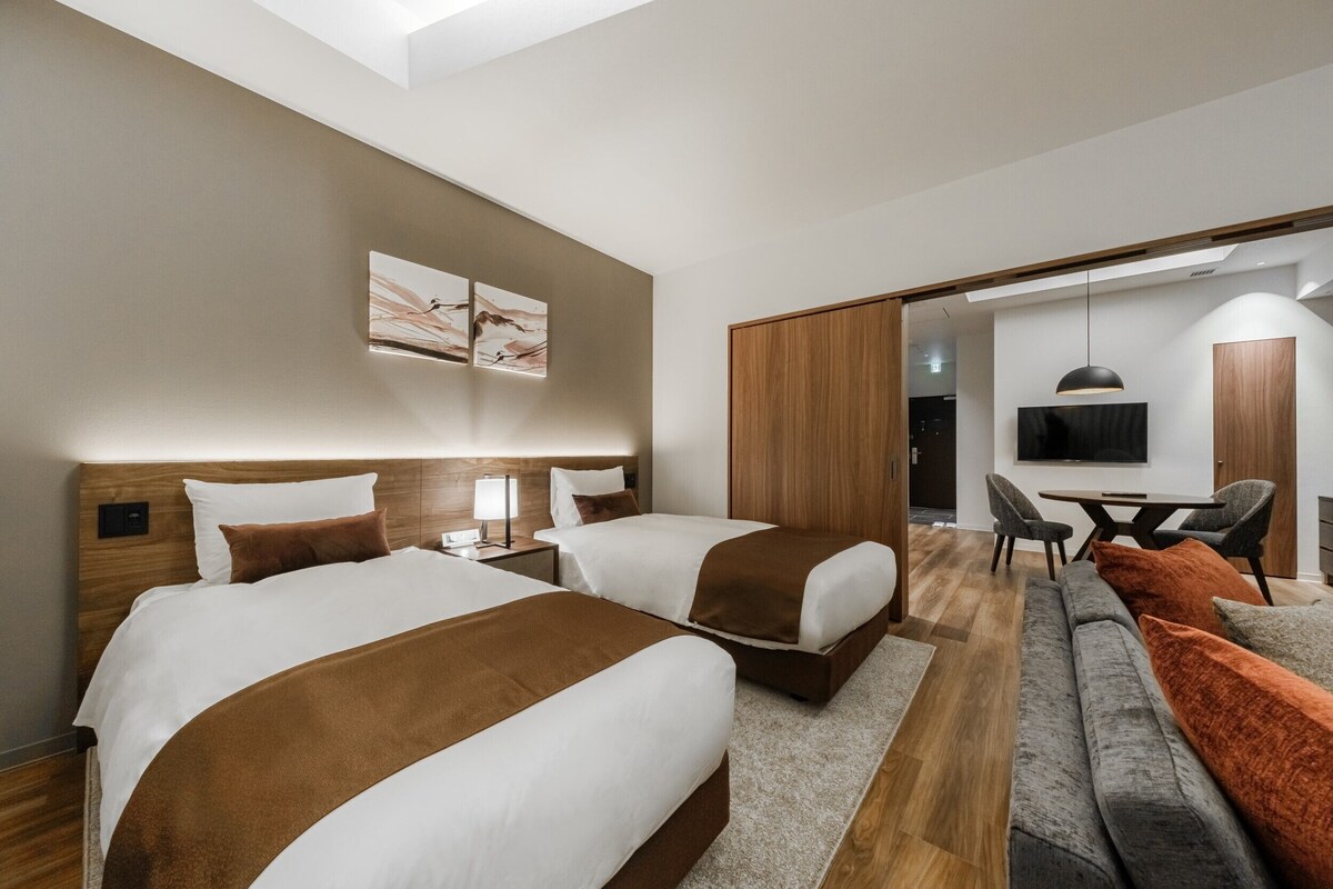 Moderni hotellihuone, jossa on kaksi sänkyä, sohva, televisio ja taideteoksia seinällä.