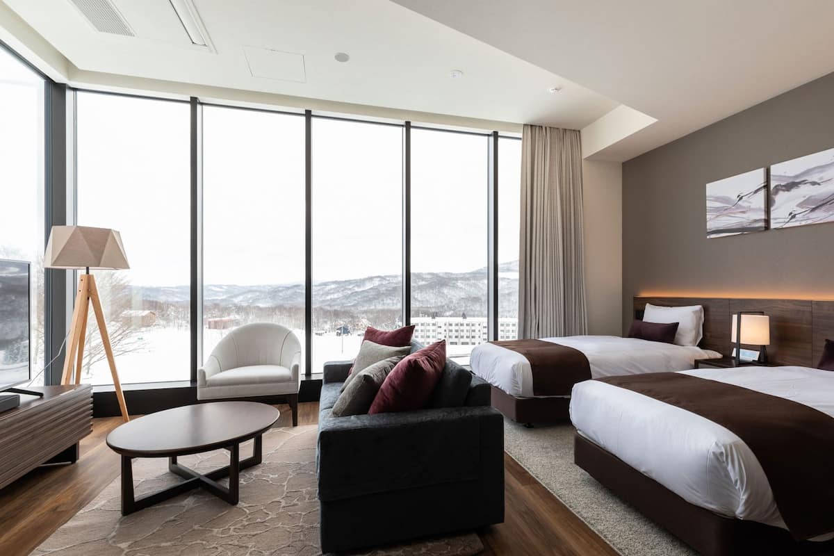 A hotel room with a large window, a sofa, a round table, two beds, and a nightstand.