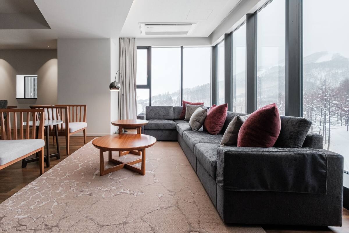 A modern living room with a grey sofa, wooden coffee table, and a view of a snowy landscape through large windows.