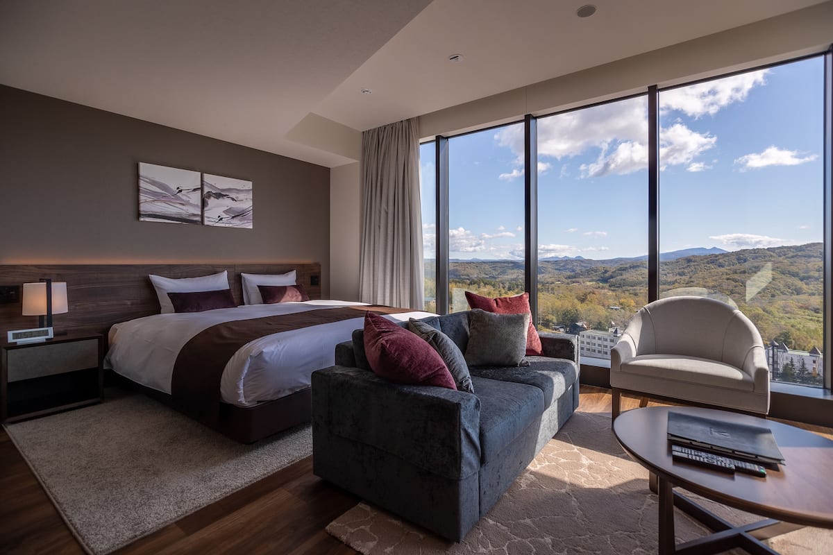 A modern hotel room with a large bed, a sofa, a chair, and a view of the mountains.