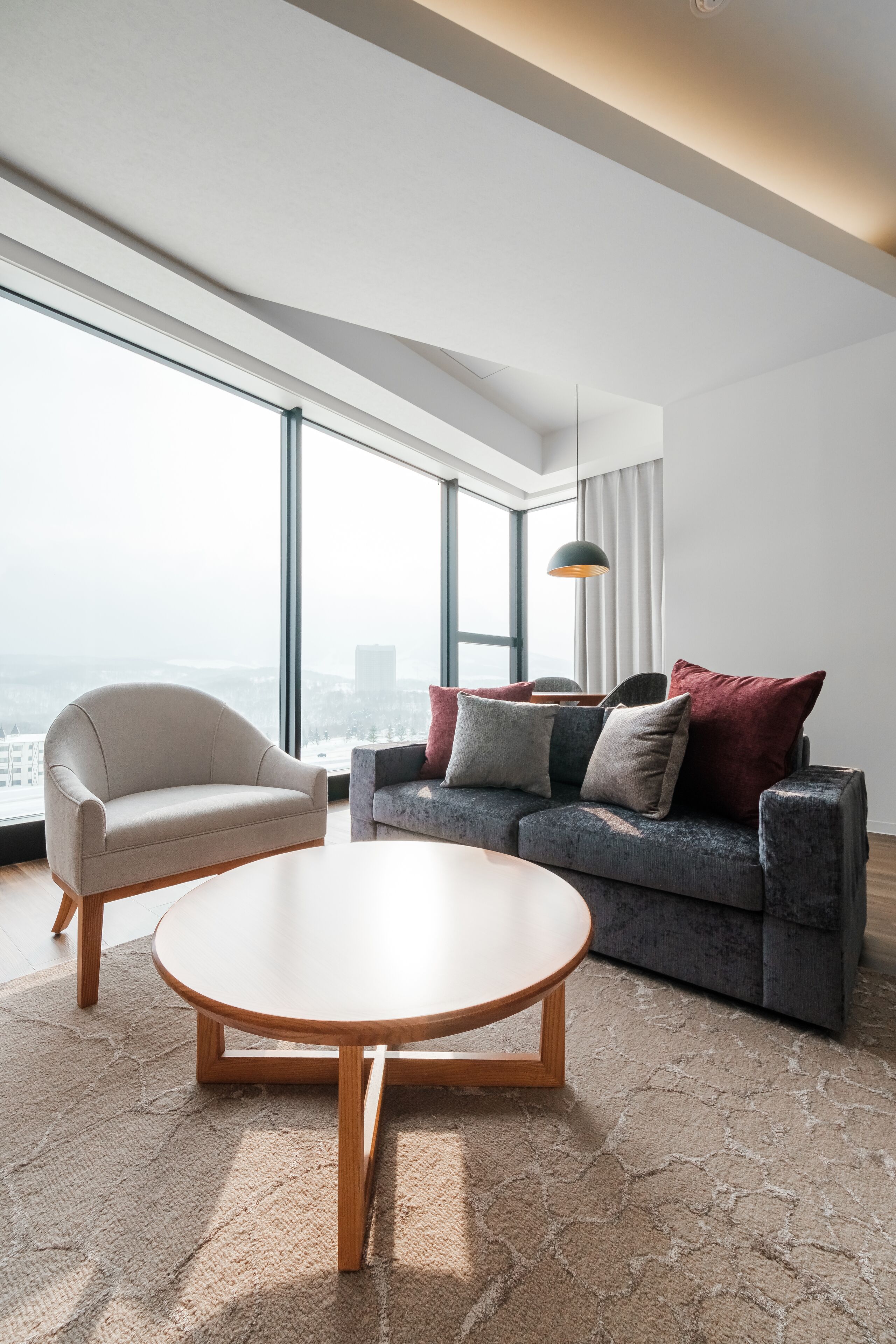 A modern living room with a sofa, armchair, and coffee table, large windows with a city view, and a pendant light.