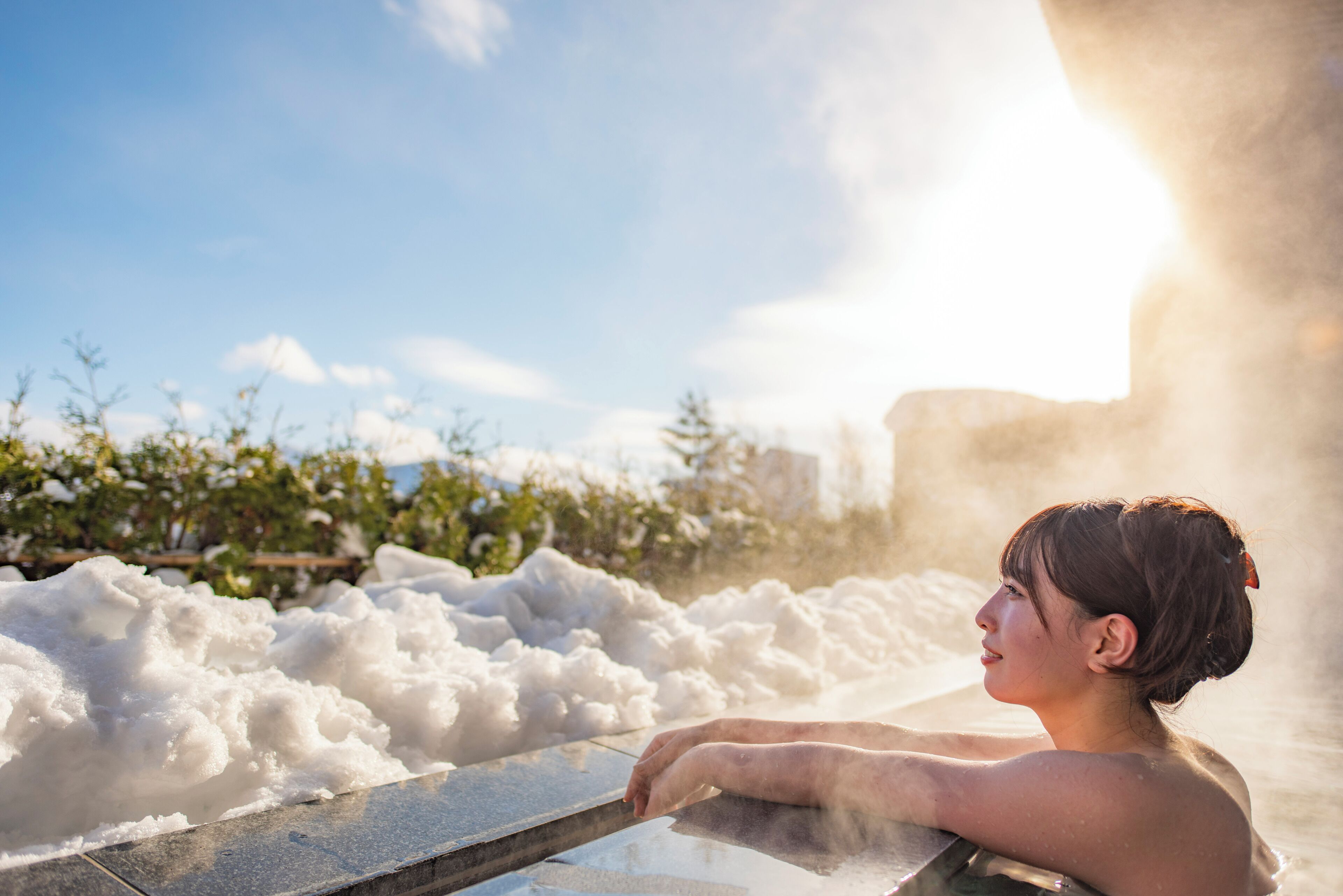 A person in a hot spring with steam rising, surrounded by snow and greenery.