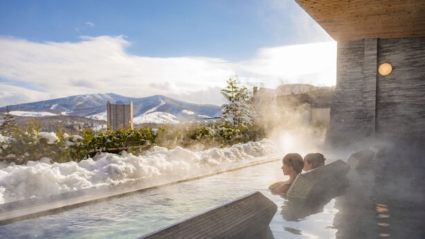 A hot spring with a view of mountains and a modern building in the background. - The Vale Rusutsu (Rusutsu)