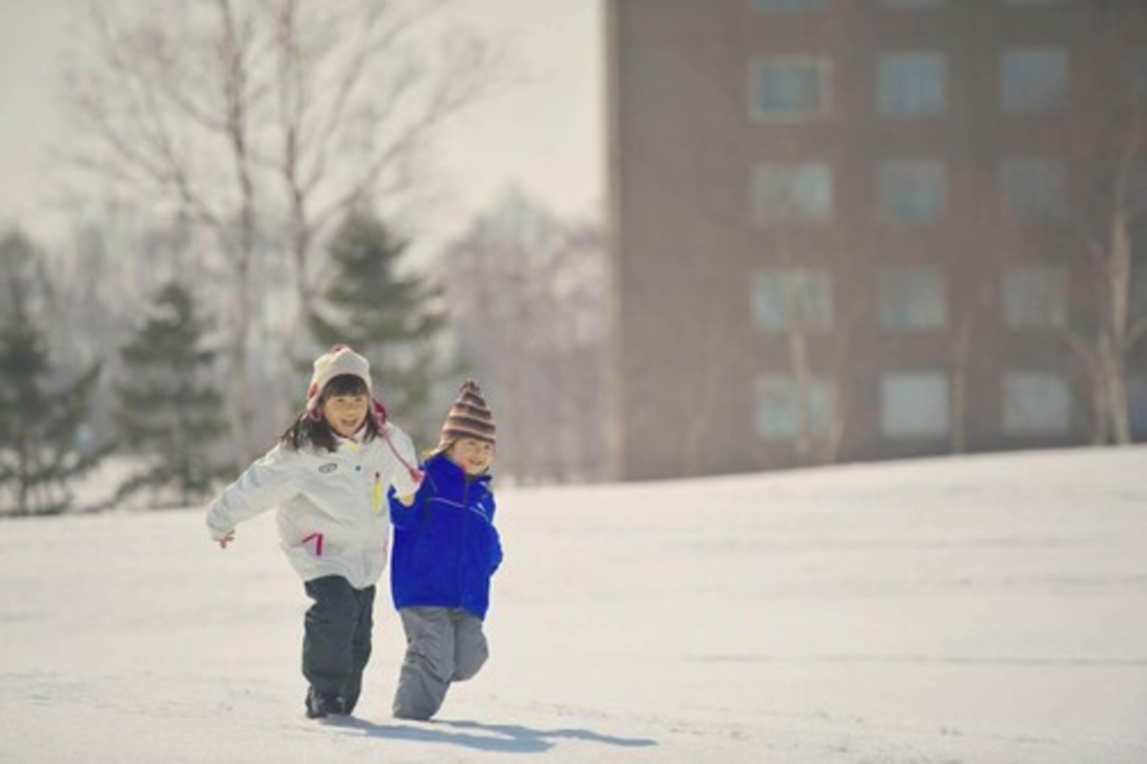 Dos niños corriendo en una zona nevada, con un edificio al fondo.