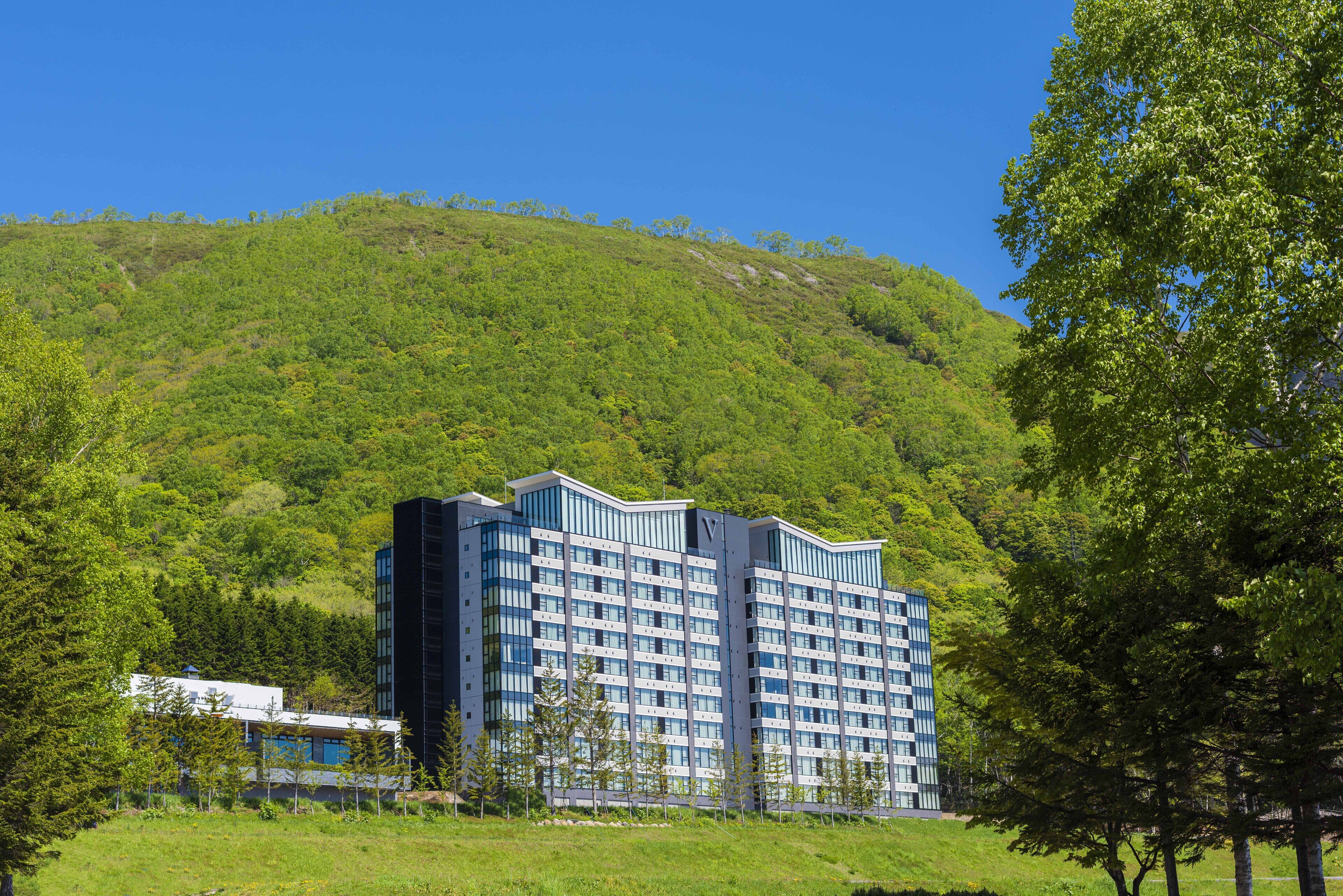 A modern hotel with a flat roof, surrounded by lush greenery and a hill in the background.