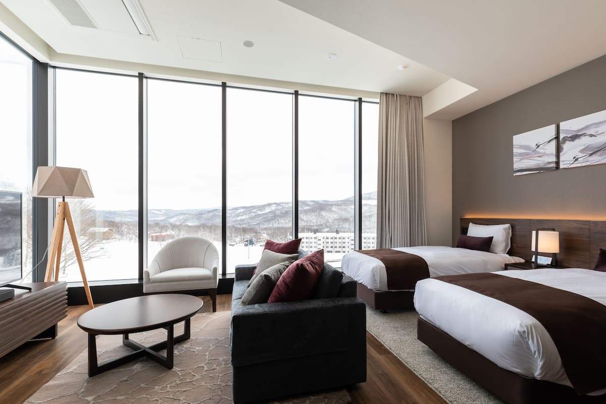 A modern hotel room with large windows, a sofa, a round coffee table, two beds, and a nightstand with a lamp.