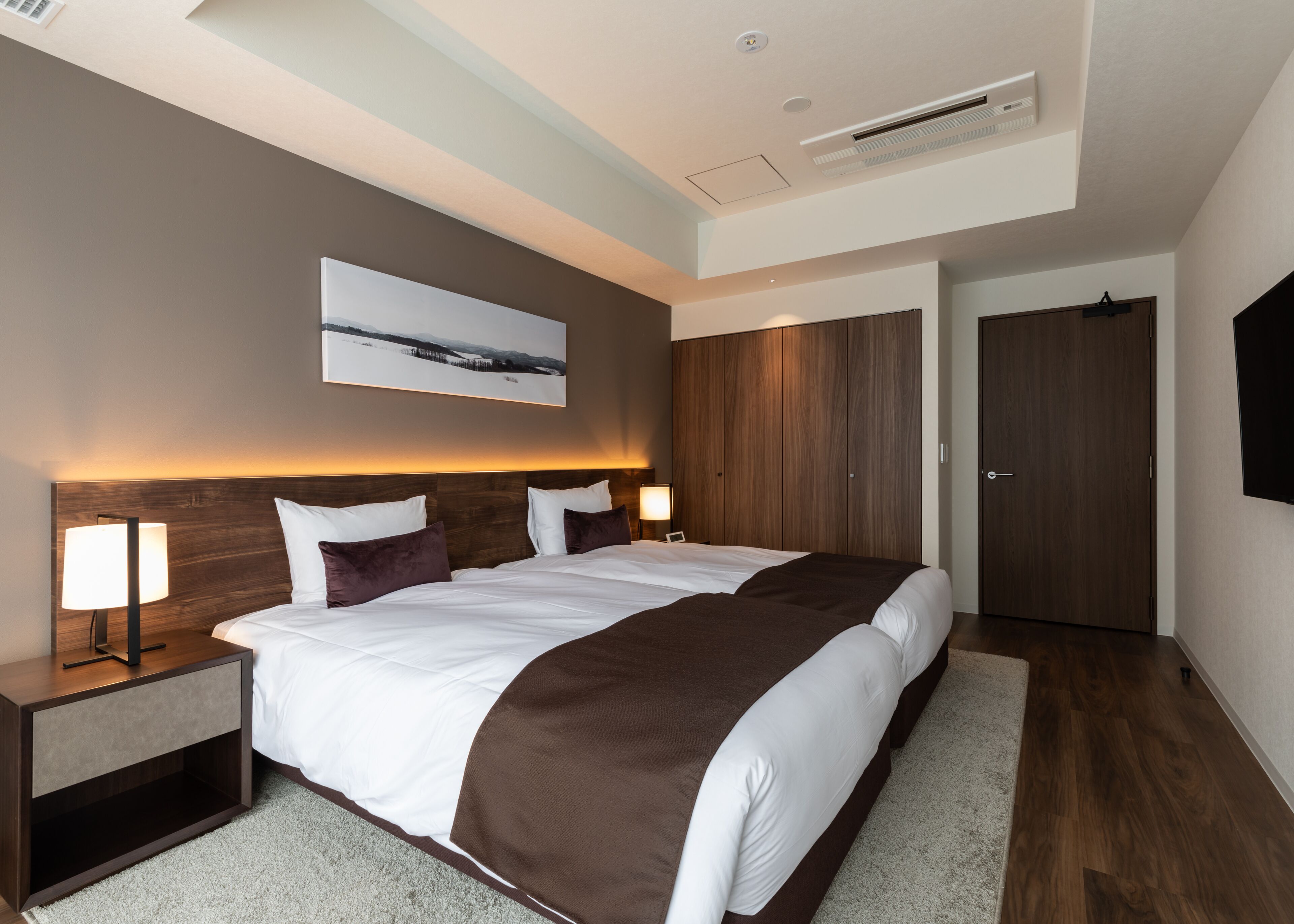 Une chambre d’hôtel moderne avec un grand lit, une table de chevet avec une lampe, un téléviseur fixé au mur et une porte.