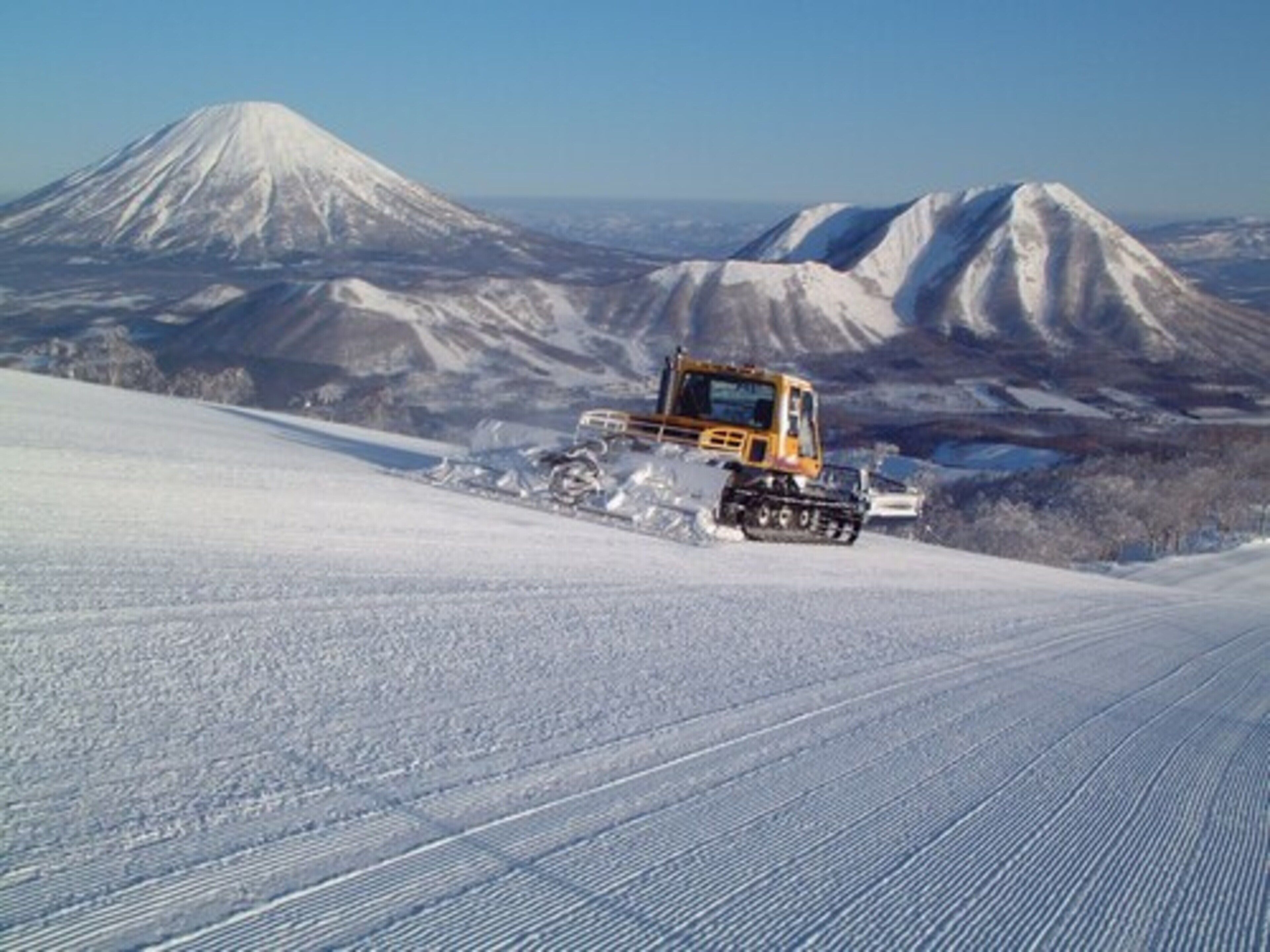 A snow-covered landscape with a large mountain in the background and a yellow construction vehicle on the snow.