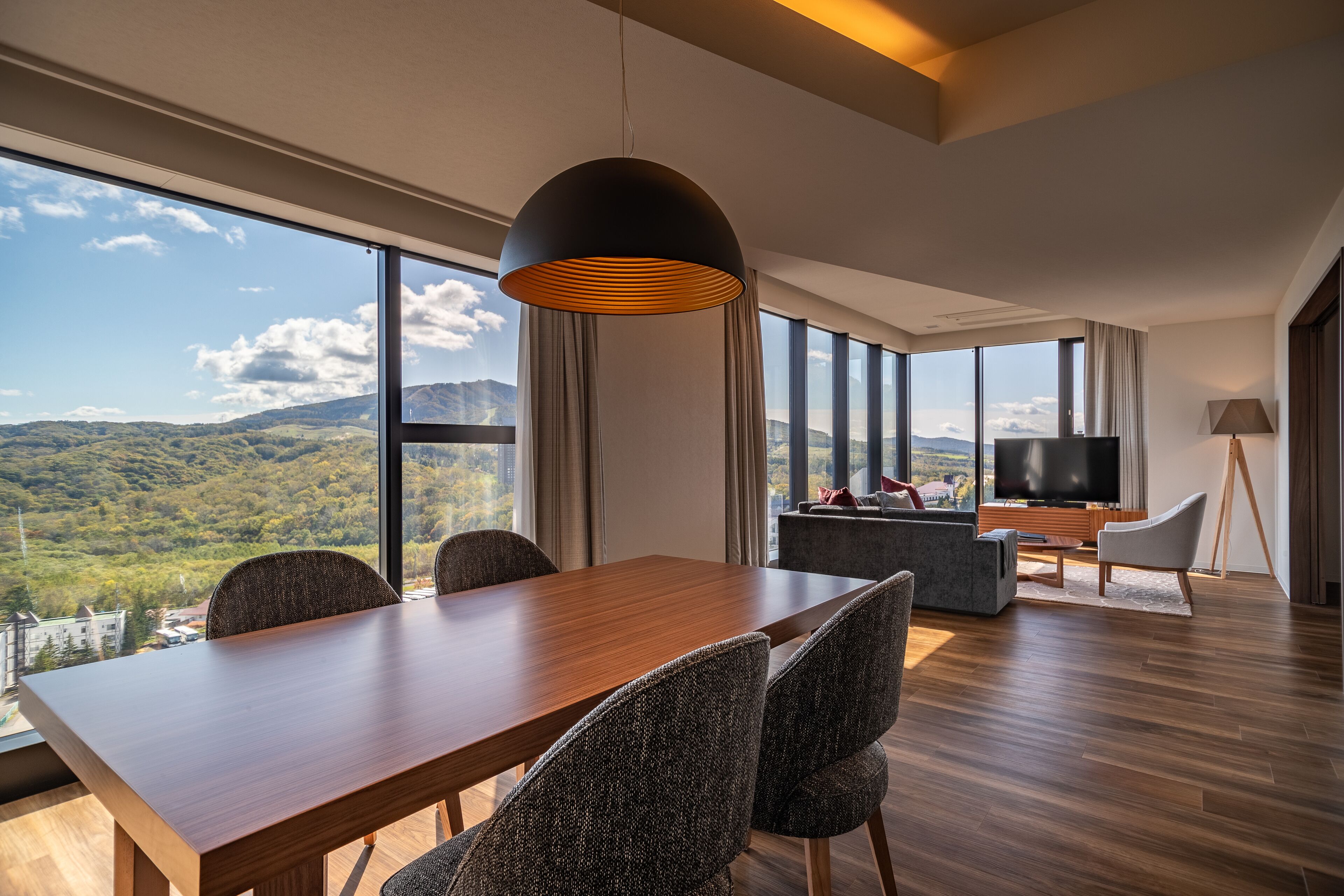 Quarto de hotel moderno com uma grande mesa de jantar, assentos confortáveis, TV de tela plana e vista panorâmica para montanhas através de amplas janelas.
