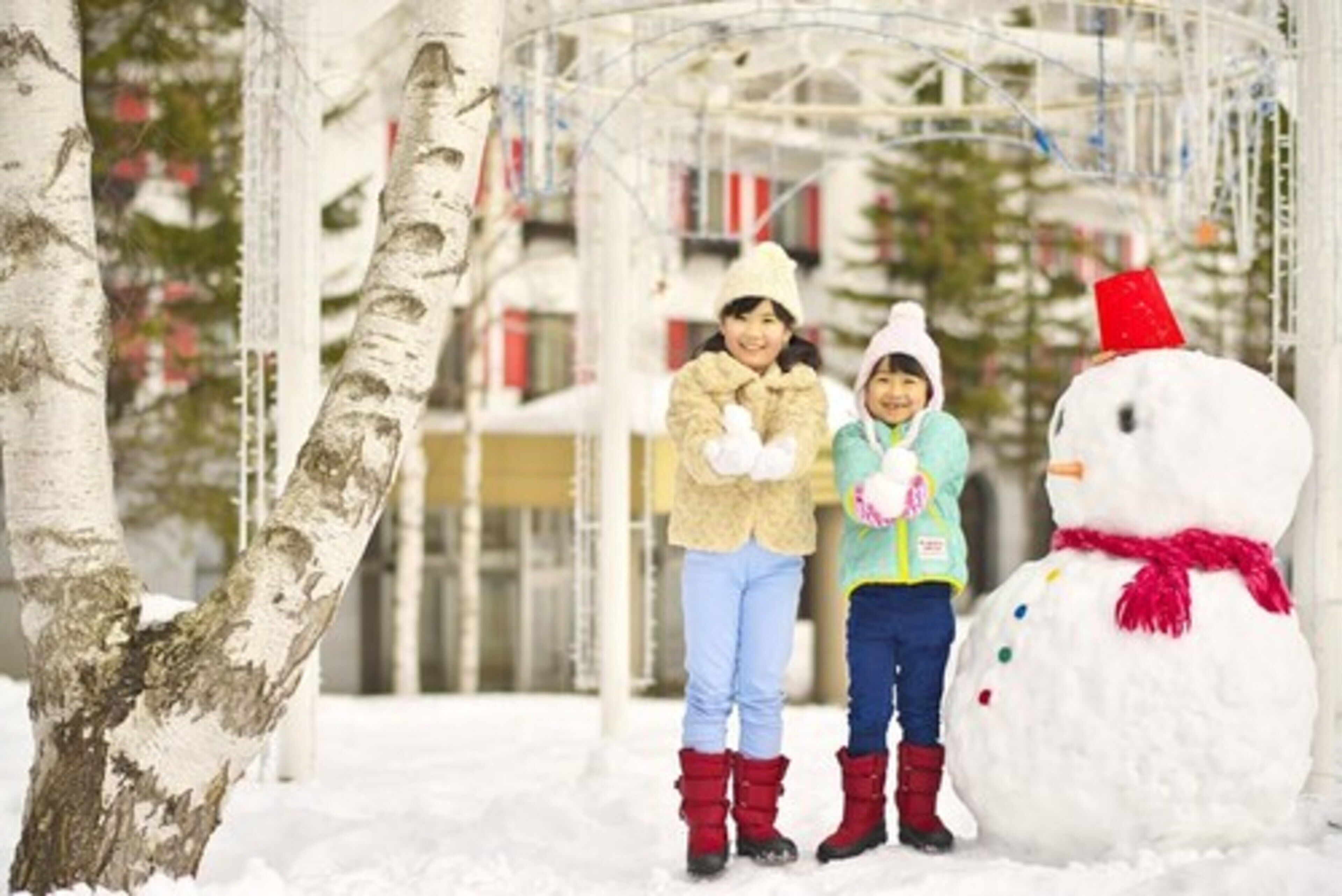 A snowman with a red hat and scarf, a person in a yellow jacket, and another in a beige jacket standing in a snowy outdoor area with trees and a metal structure in the background.