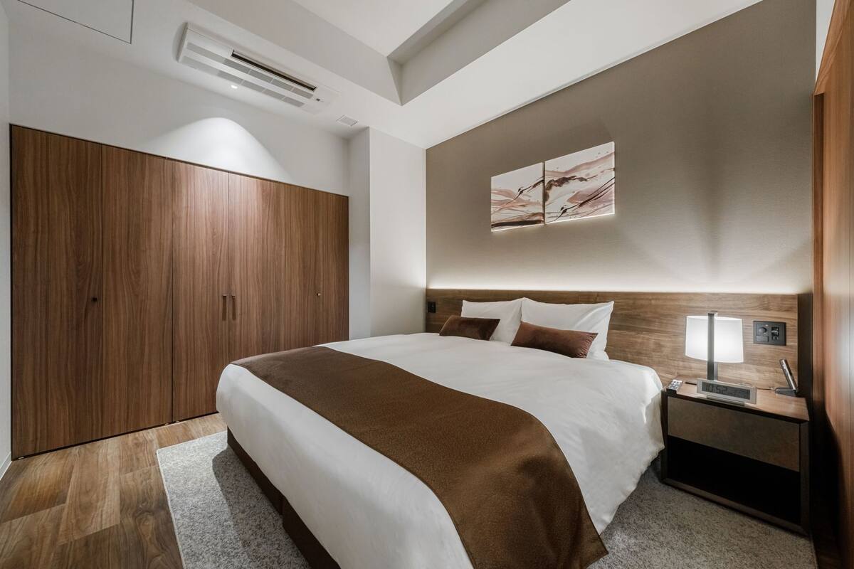 A modern hotel room with a large bed, wooden wardrobe, and a nightstand with a lamp and clock.