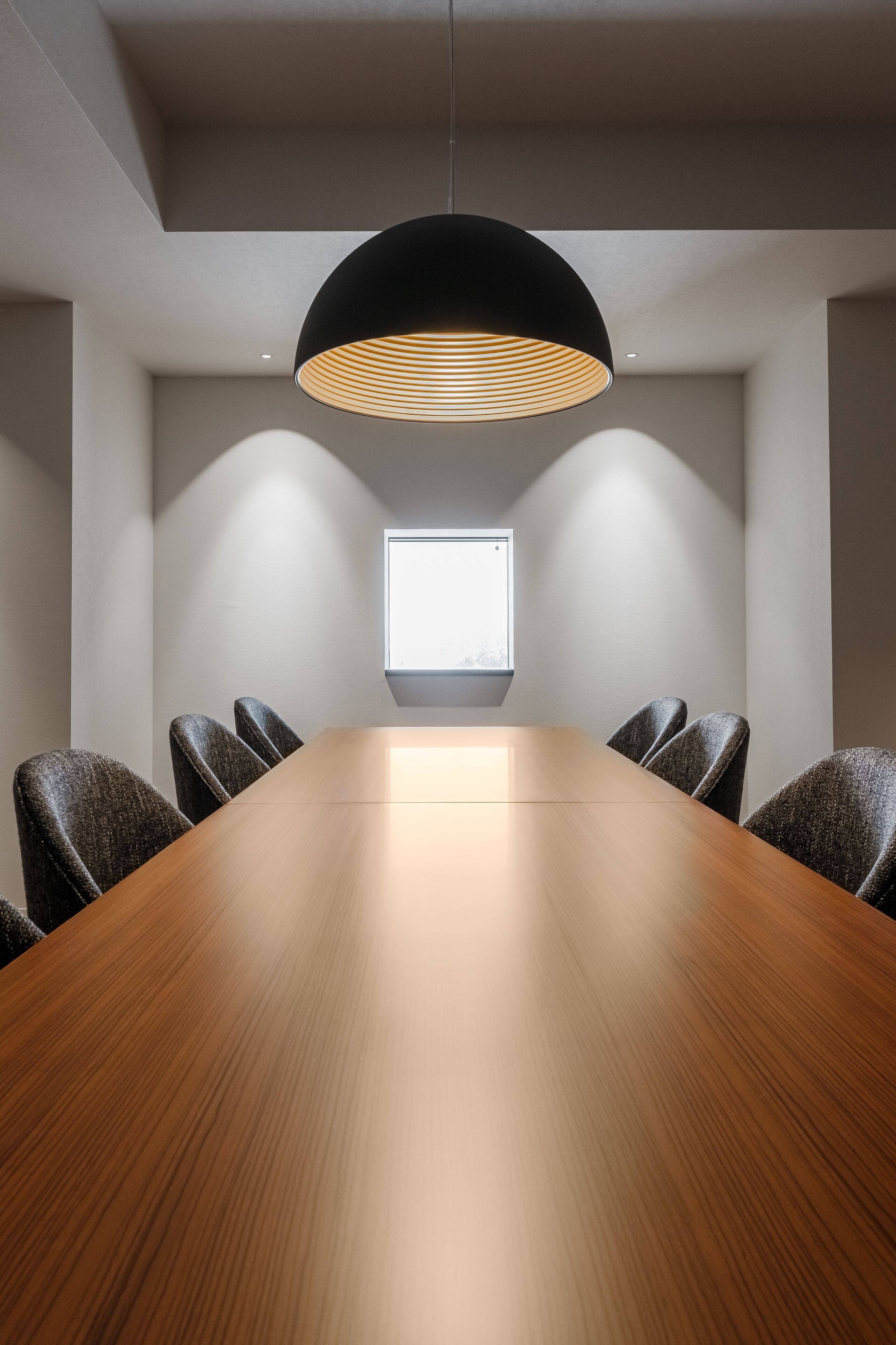 A conference room with a long wooden table, six chairs, a black pendant light, and a window with natural light.
