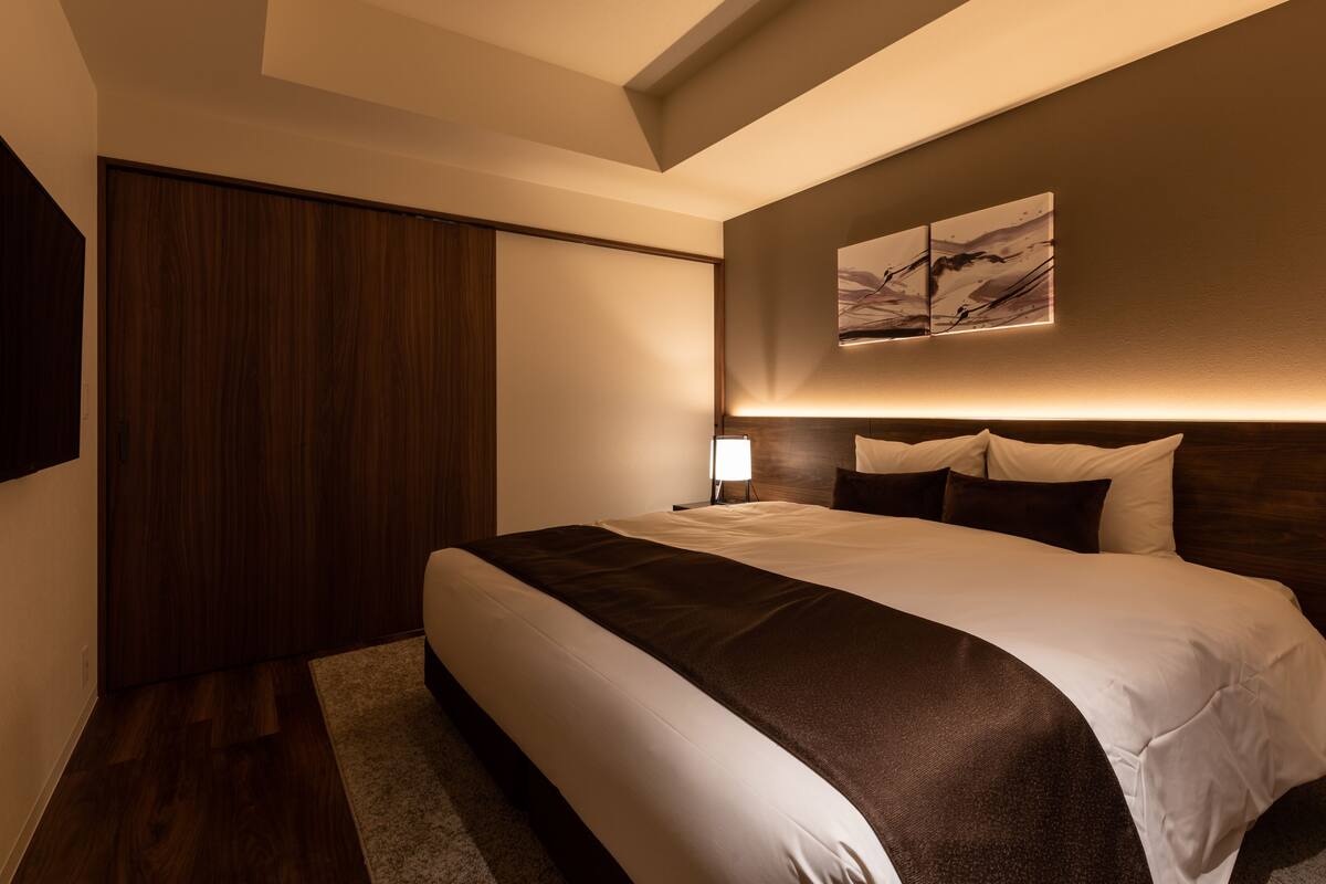 A modern hotel room with a large bed, a wooden wardrobe, a wall-mounted artwork, and a ceiling light.