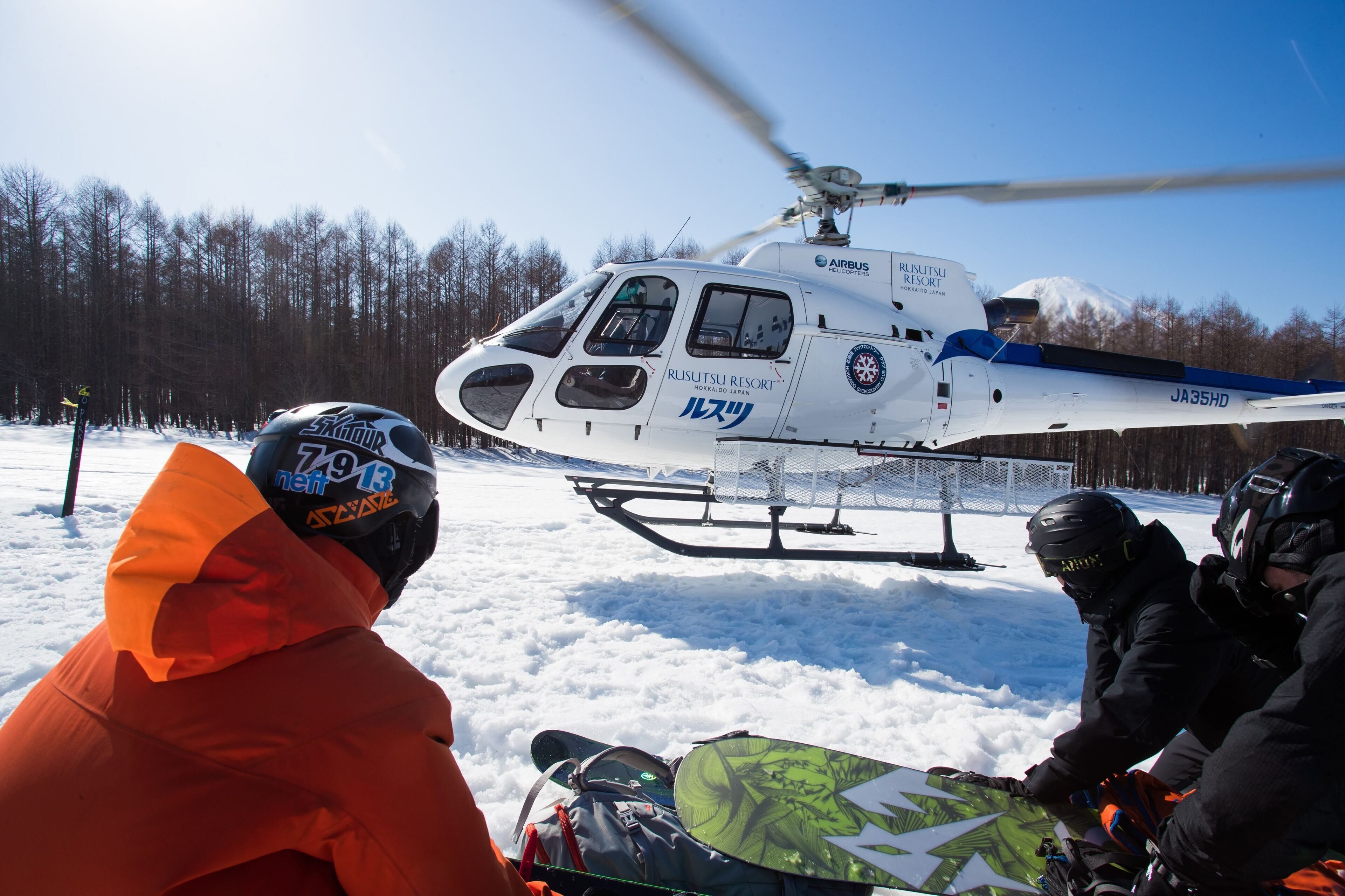 A helicopter with "Rusutsu Resort" and "JA35HD" on its side hovering over a snowy landscape with trees in the background.