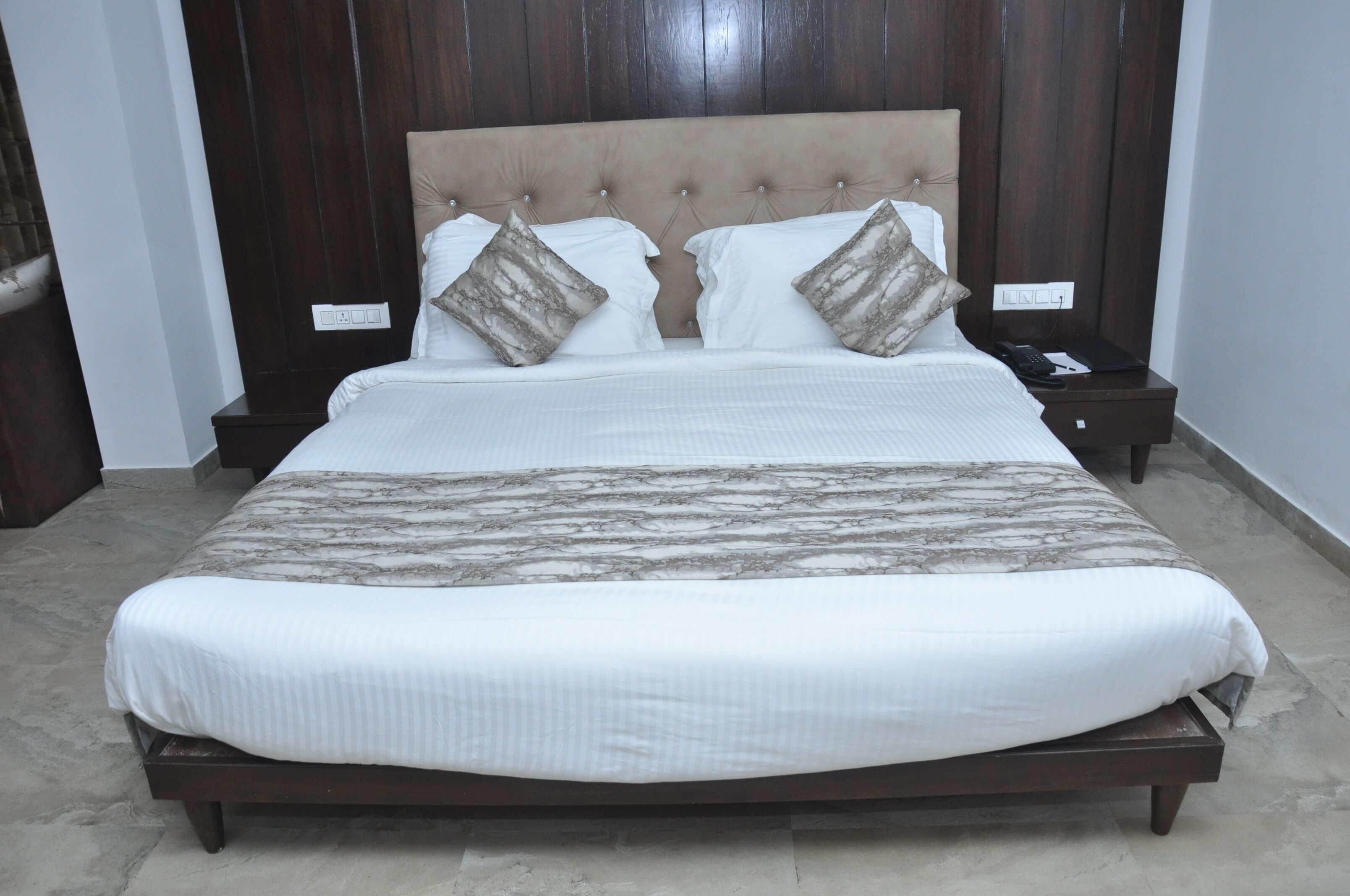 A neatly made bed with a wooden headboard, two nightstands with a phone and a book, and a marble-patterned bedspread.