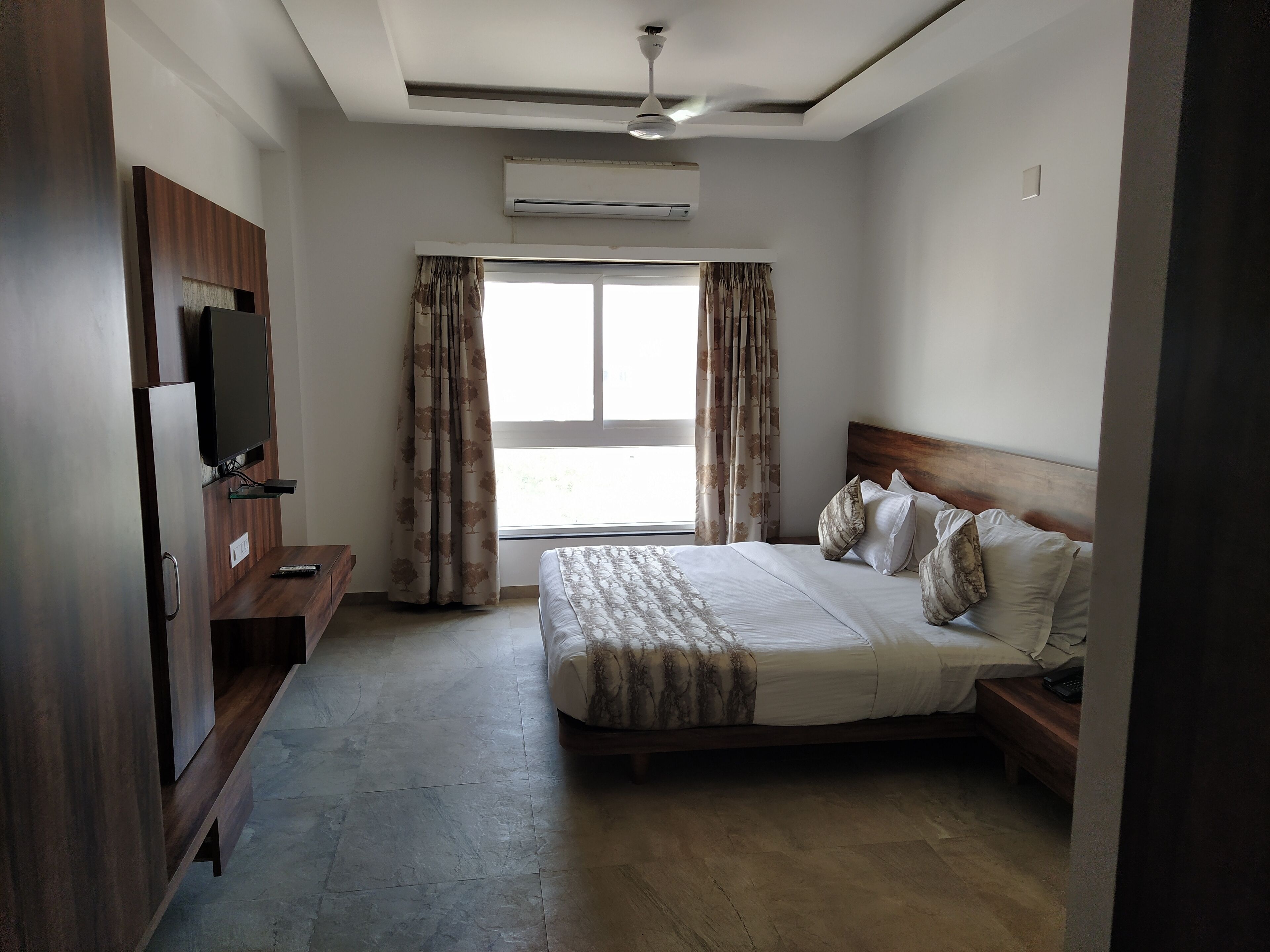 A hotel room with a bed, a TV, a desk, and a window with curtains.