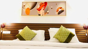 Quarto de hotel com cabeceira de madeira, duas almofadas verdes estampadas e uma pintura abstrata emoldurada acima da cama.