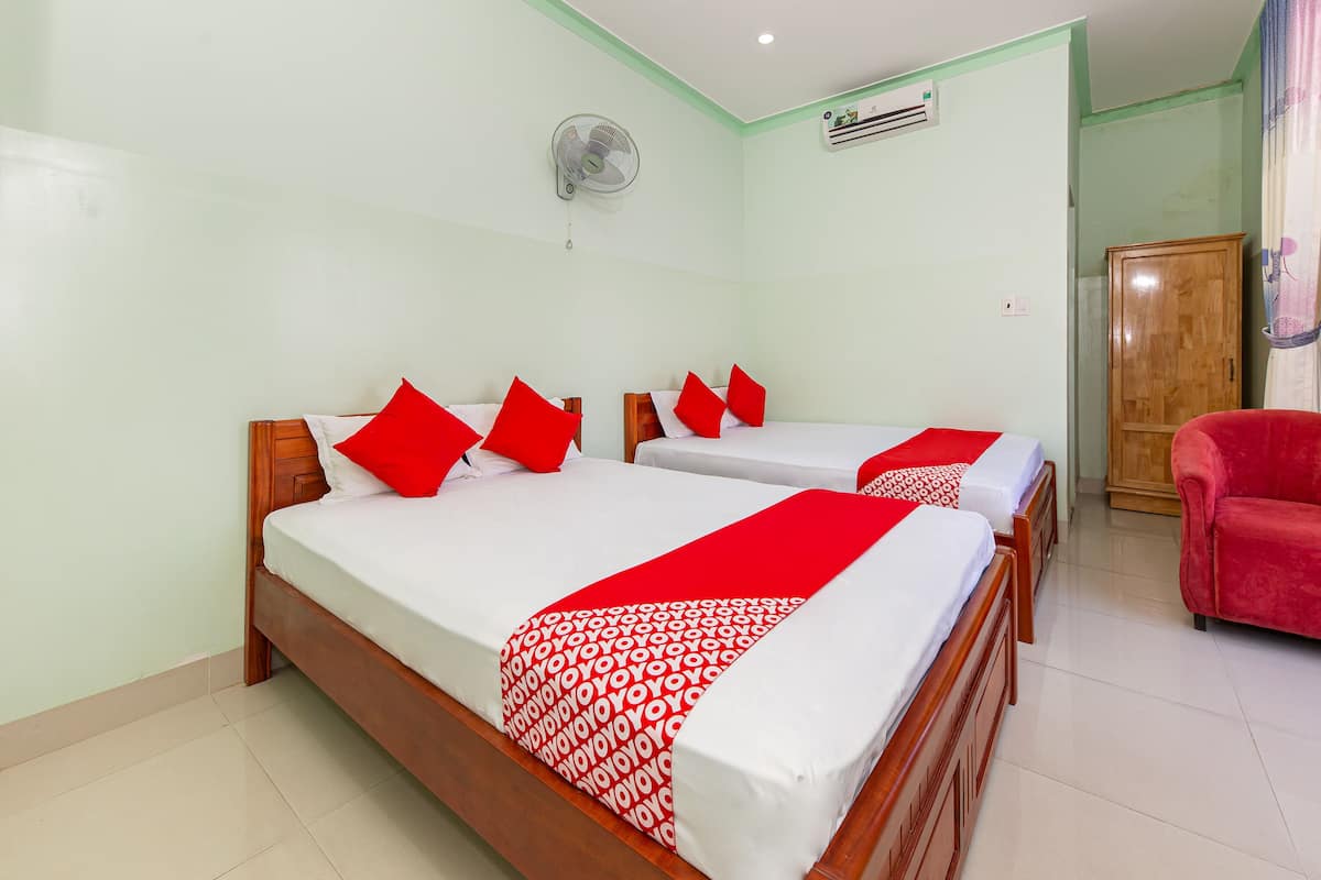Une chambre d’hôtel avec deux lits, chacun agrémenté d’oreillers rouges et d’une couvre-lit à motifs rouges et blancs. On y trouve également une armoire en bois, une chaise rouge et un ventilateur de plafond.