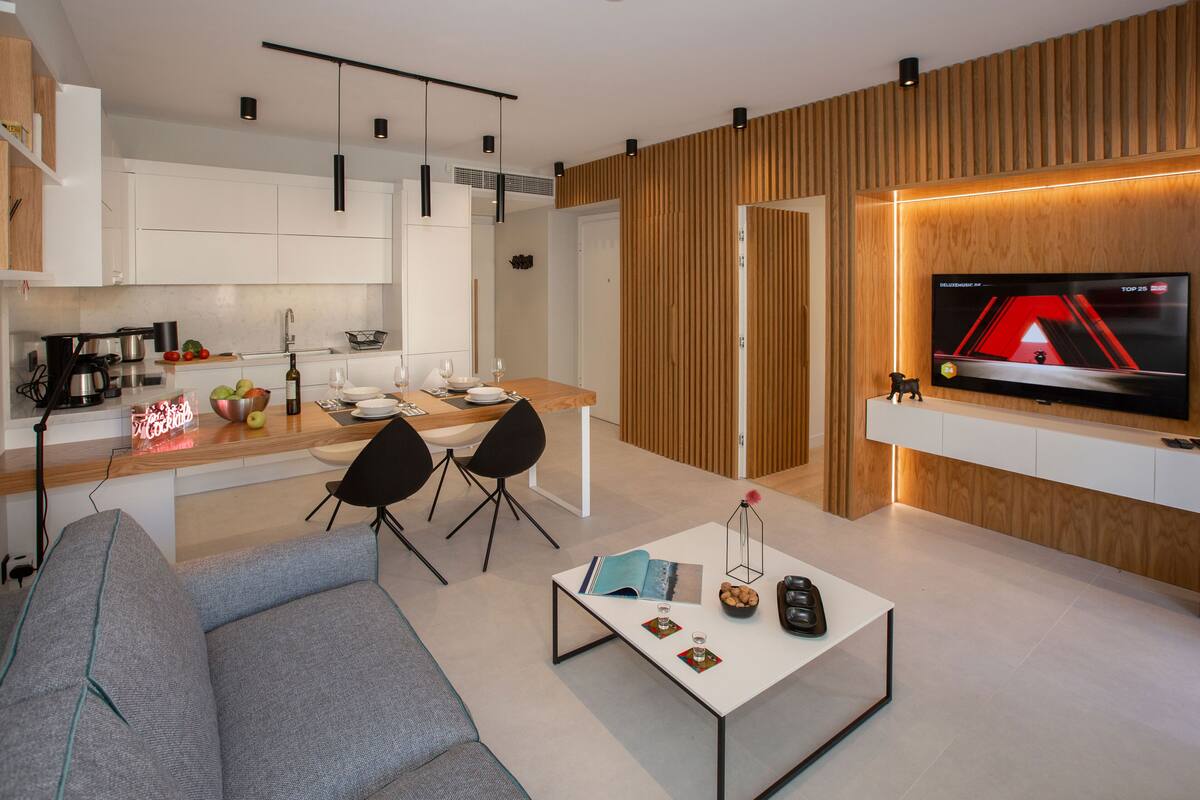 A modern living room with a grey sofa, a coffee table with books and a candle, a TV mounted on a wooden wall, and a kitchen area with white cabinets and a wooden countertop.