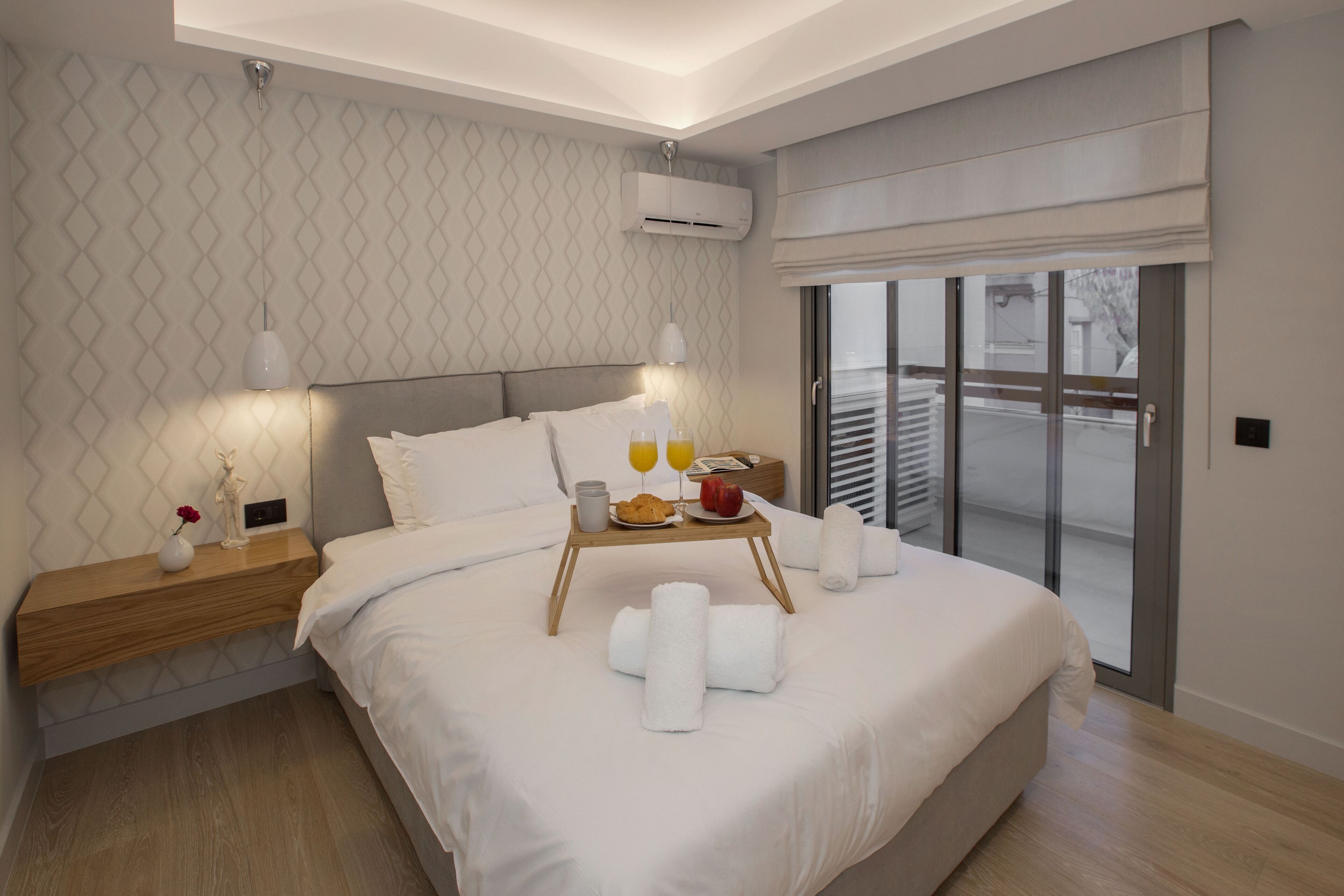 A modern hotel room with a large bed, a bedside table with a lamp, a tray with towels and breakfast items, and a sliding glass door to the outside.