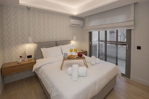Quarto de hotel moderno com uma cama grande, uma mesa de cabeceira com abajur, um suporte com toalhas e itens de café da manhã, e vista para o exterior por uma porta deslizante.