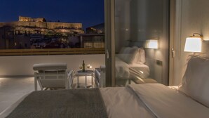 A hotel room with a bed, a small table with a bottle and glass, a chair, and a view of ancient ruins at night. - Acropolis Golden Suites by GHH (Athens)