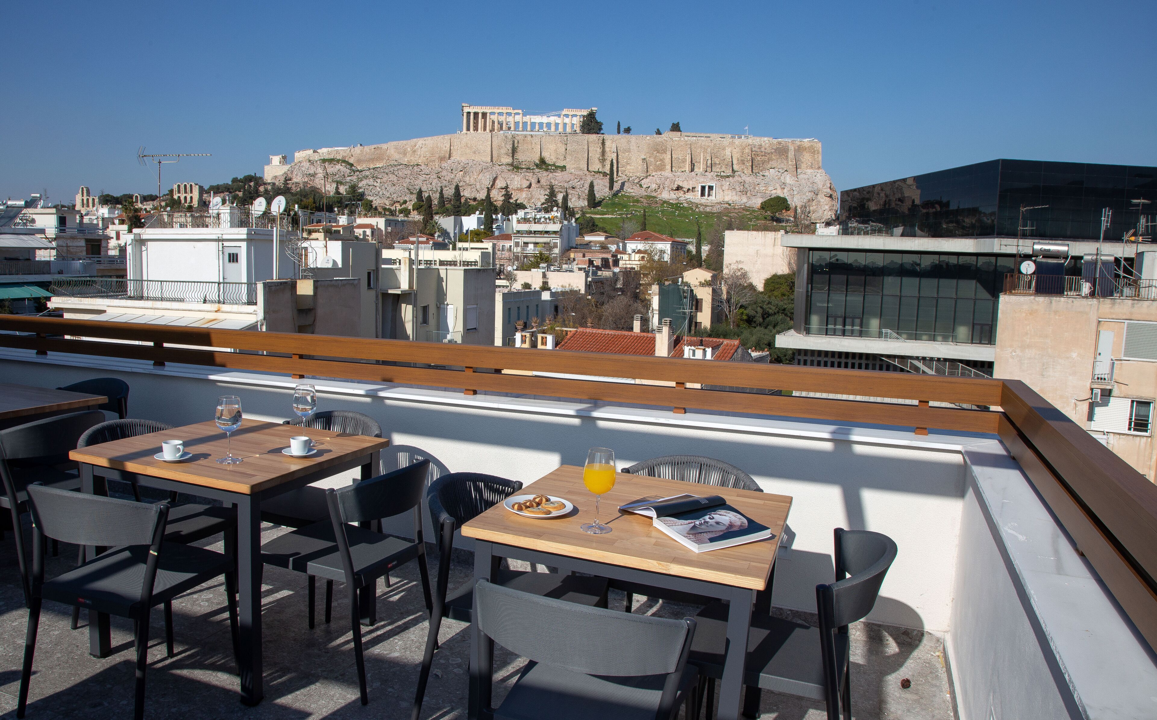 Una terraza en la azotea con mesas y sillas, con vistas a un paisaje urbano histórico que incluye ruinas antiguas y edificios modernos.