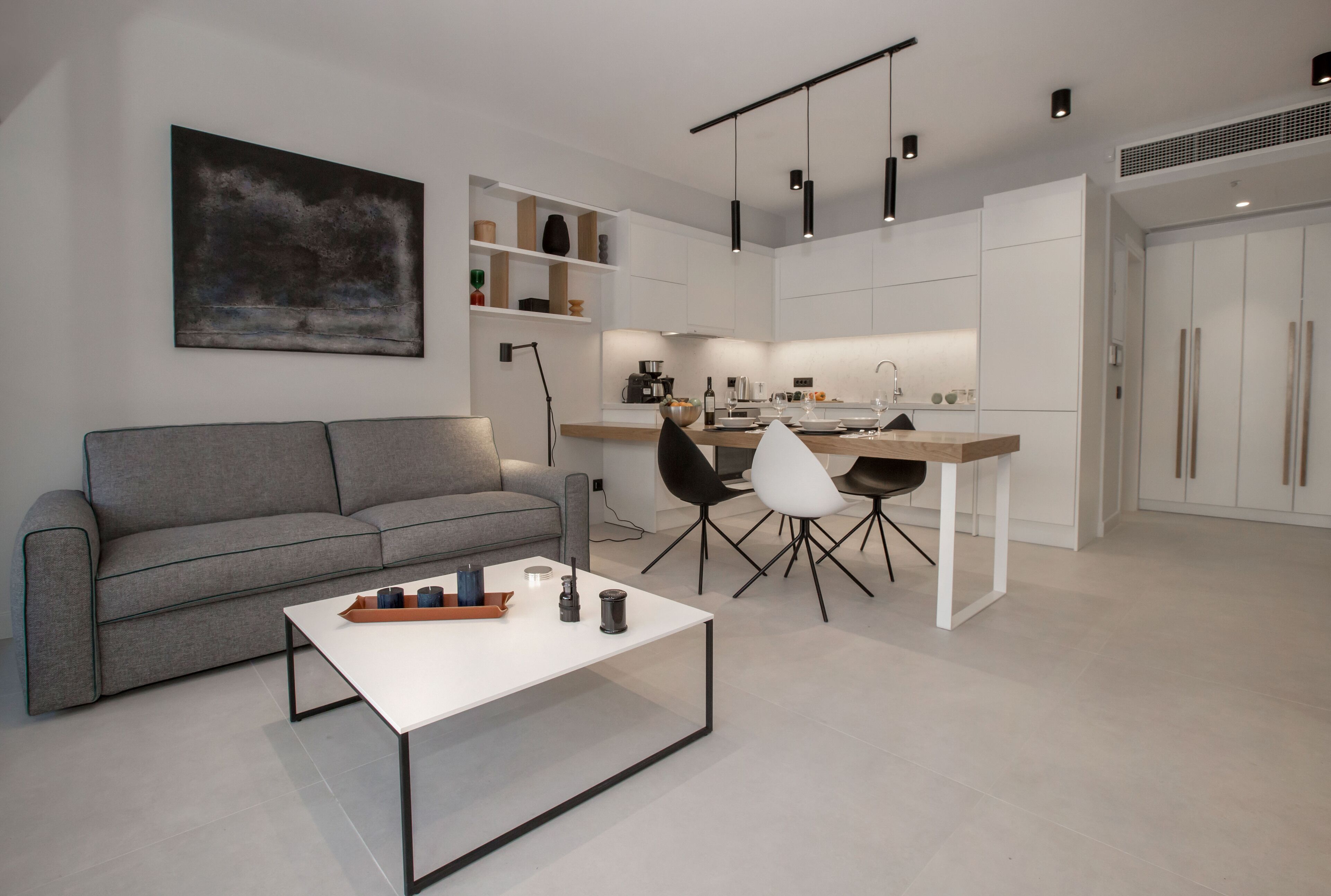 A modern living room with a grey sofa, a white coffee table, and black chairs. There is a kitchen area with white cabinets and a dining table.
