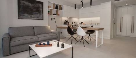 A modern living room with a grey sofa, a white coffee table, and black chairs. There is a kitchen area with white cabinets and a dining table.