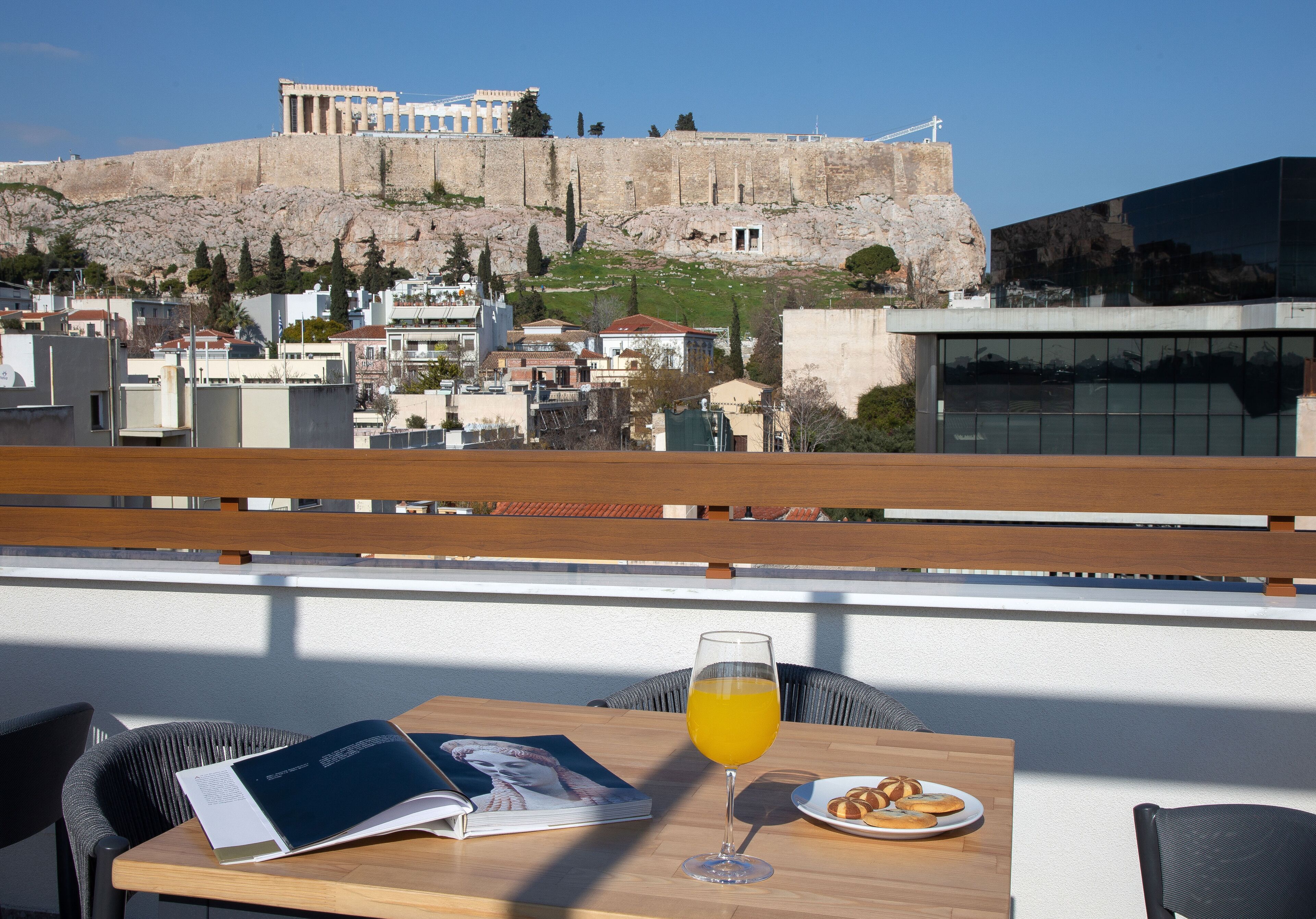 Un tavolo con un libro, un bicchiere di succo d'arancia e un piatto di dolci, con vista su una città e rovine antiche sullo sfondo.