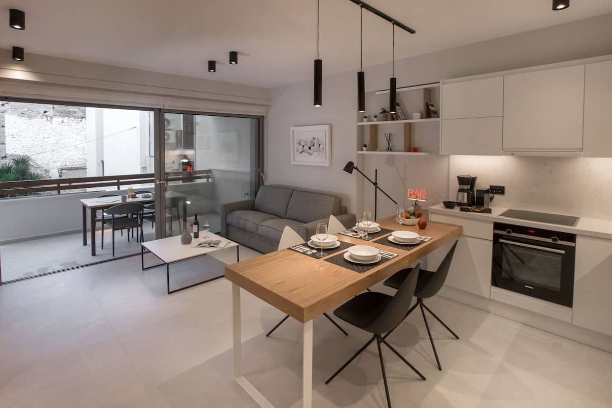 A modern kitchen with white cabinets, a dining area with a wooden table, and a balcony with a view of the outside.