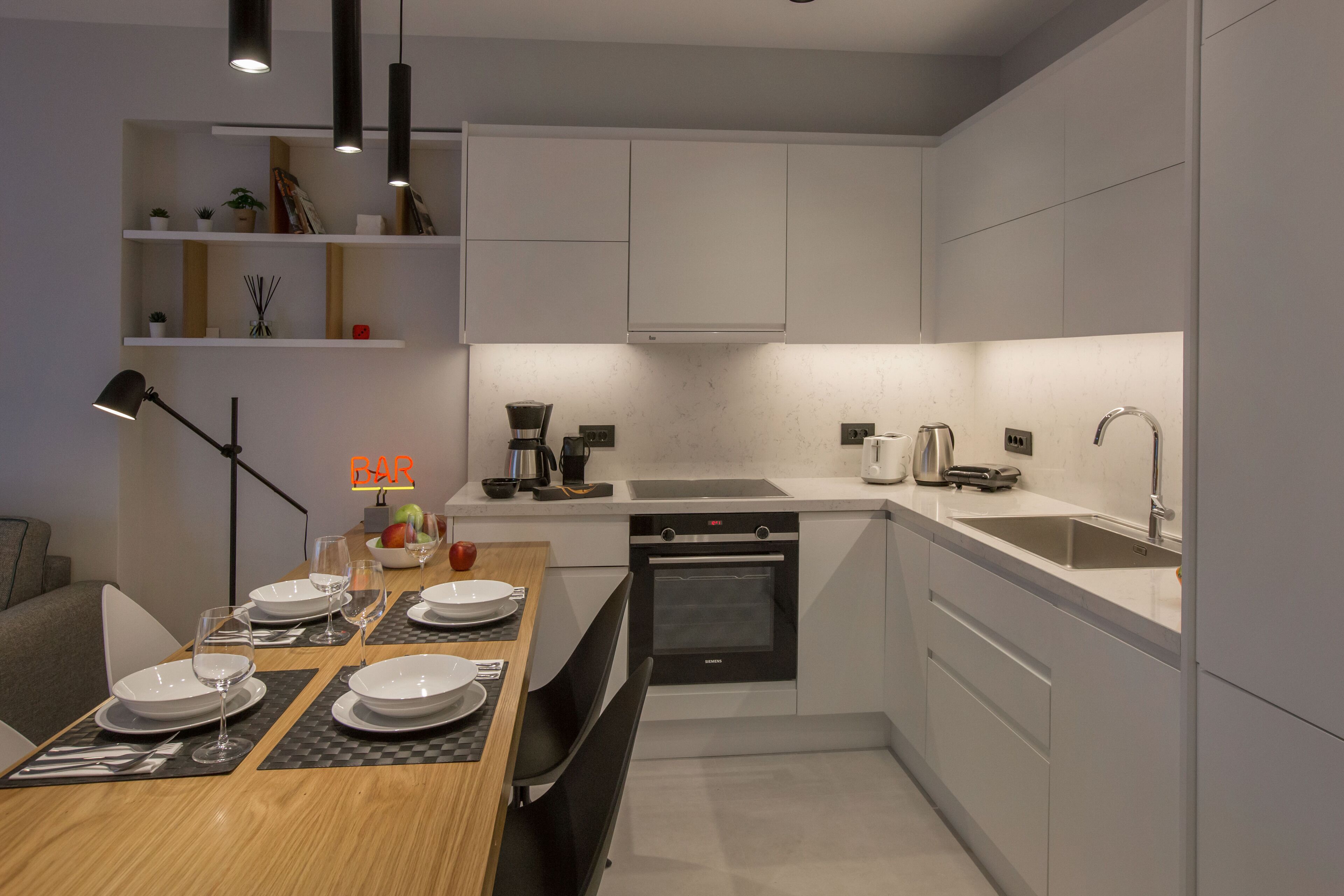 A modern kitchen with white cabinets, a built-in oven, a sink, and a dining area with a table set for four.
