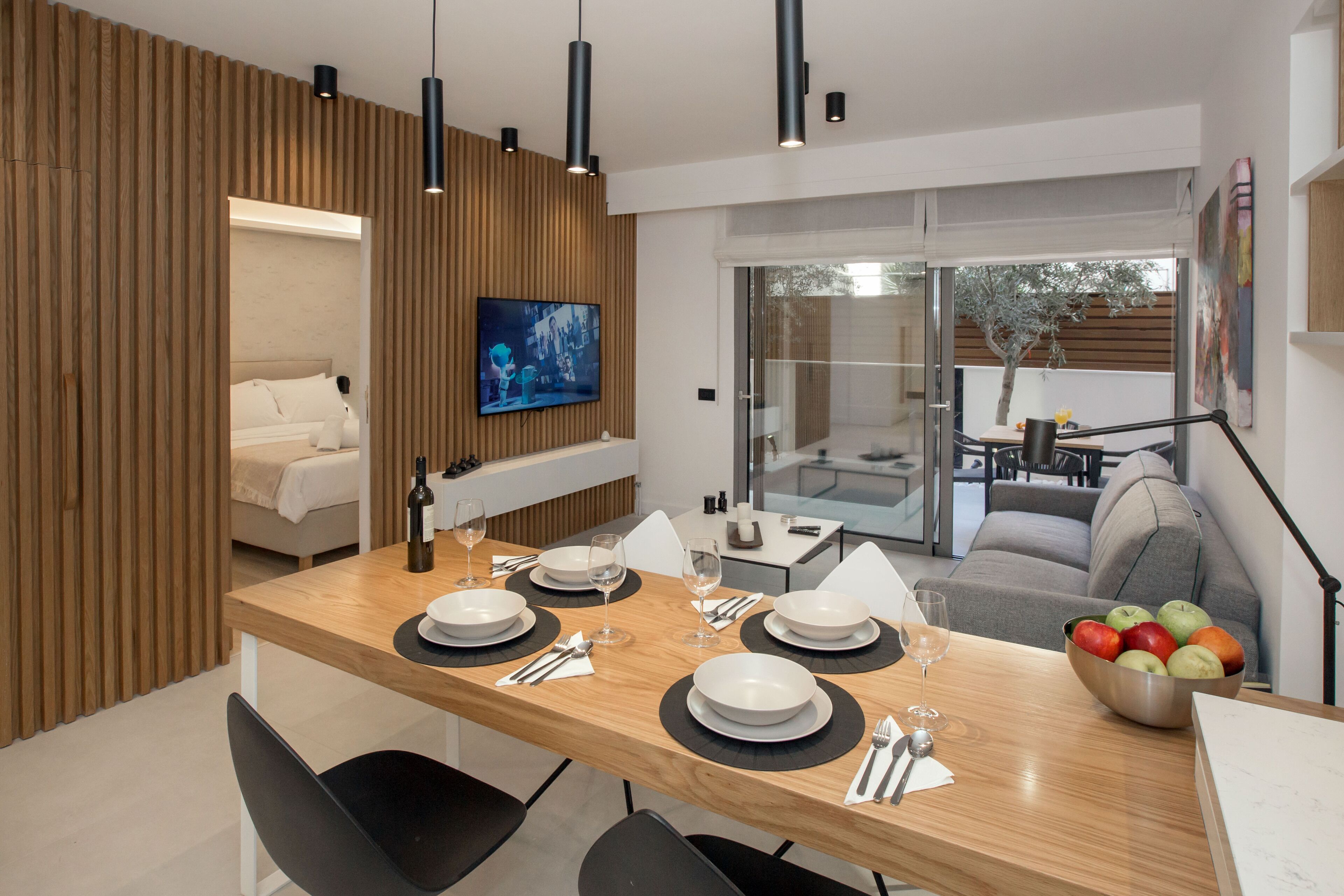 A modern dining area with a wooden table set for four, a television mounted on the wall, and a view of the outdoor patio.