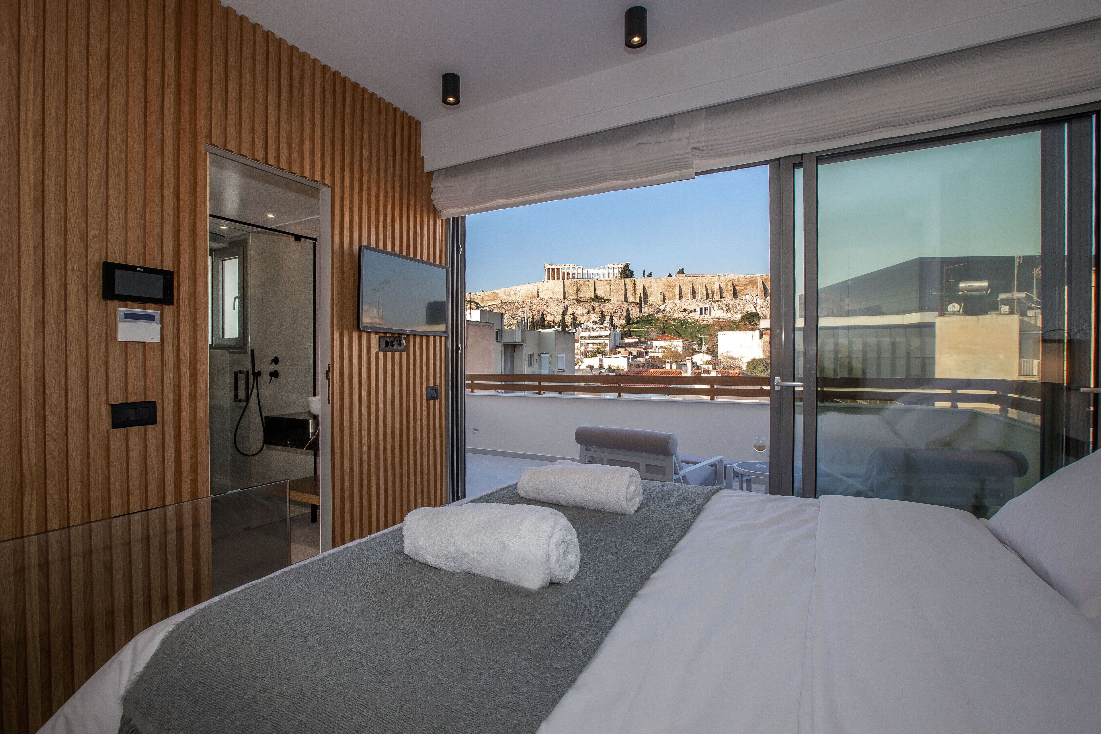 A modern hotel room with a wooden panel wall, a large bed with white linens, a glass shower enclosure, and a balcony with a view of a historic hill.