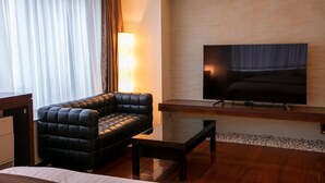 Une chambre d’hôtel moderne dotée d’un grand téléviseur à écran plat fixé à un mur en briques, d’un canapé en cuir noir, d’une table basse en bois et d’un lit recouvert d’une couette blanche.