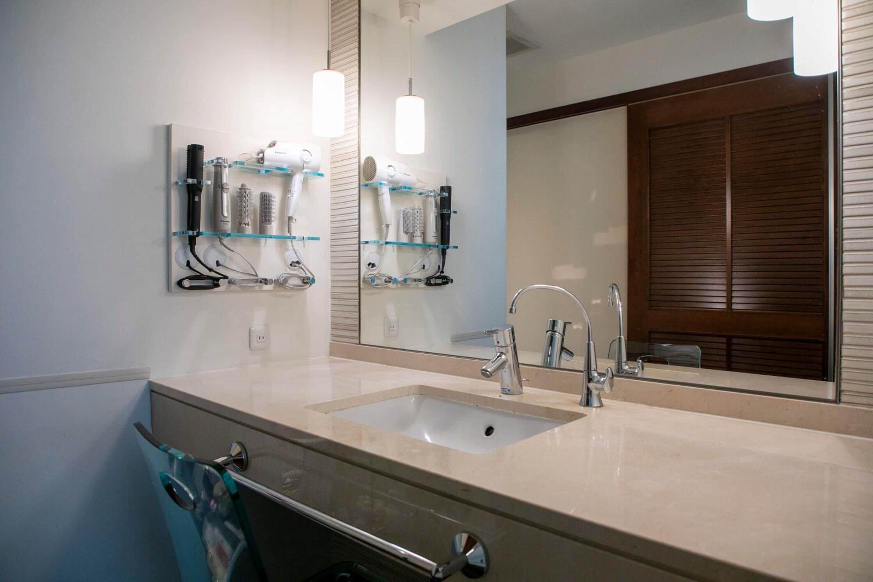 A bathroom with a double sink, a mirror, and a glass shelf with hairdryers.