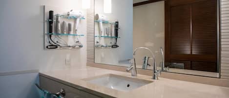 A bathroom with a double sink, a mirror, and a glass shelf with hairdryers.