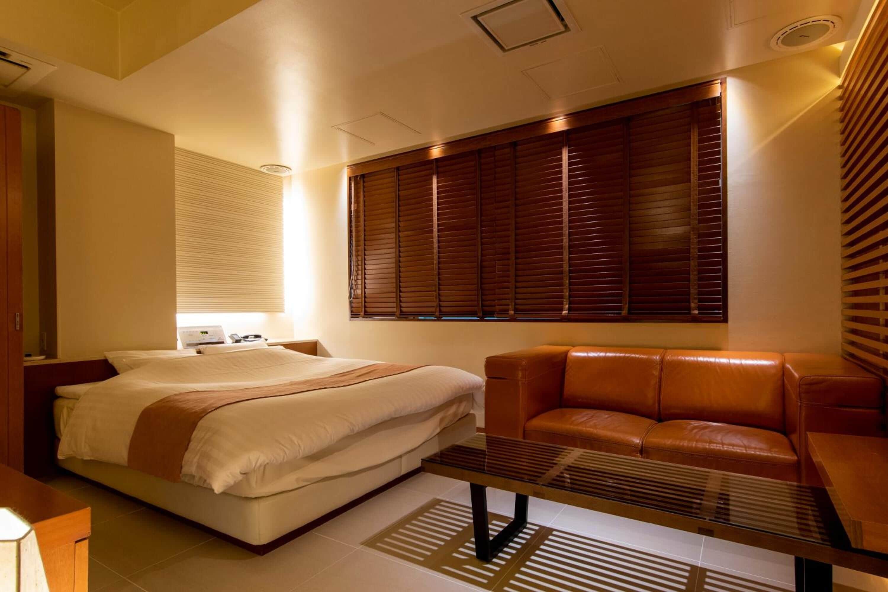 Une chambre d’hôtel moderne, équipée d’un lit, d’un canapé en cuir, d’une table basse en verre et d’une grande fenêtre avec des stores.
